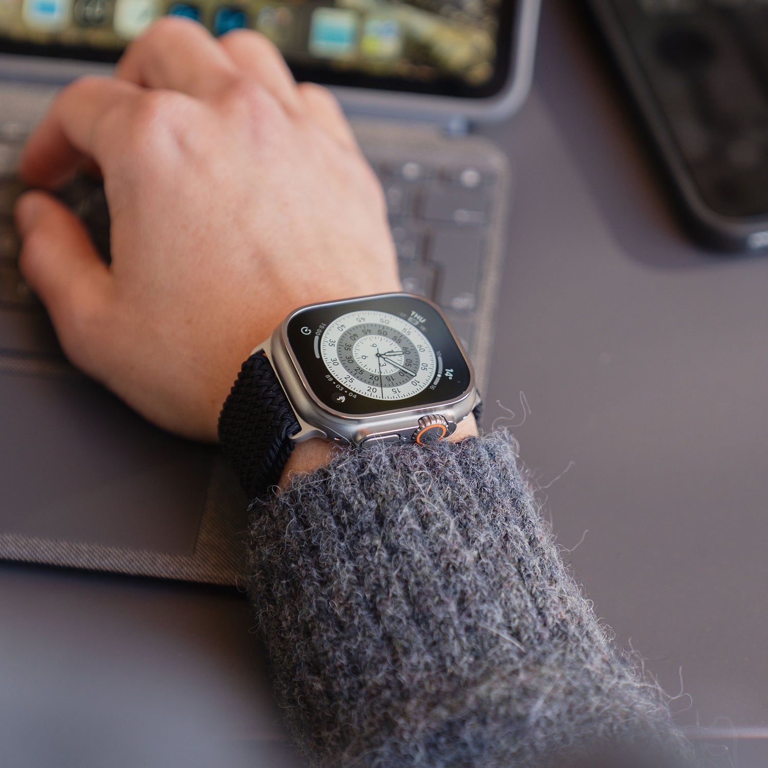 Eine Person, die das Arktisband „Geflochtenes Apple Watch Ultra Armband ‚Casual‘“ mit Magnetverschluss trägt, tippt auf einer Tastatur, während im Hintergrund ein Smartphone und ein Laptop zu sehen sind.