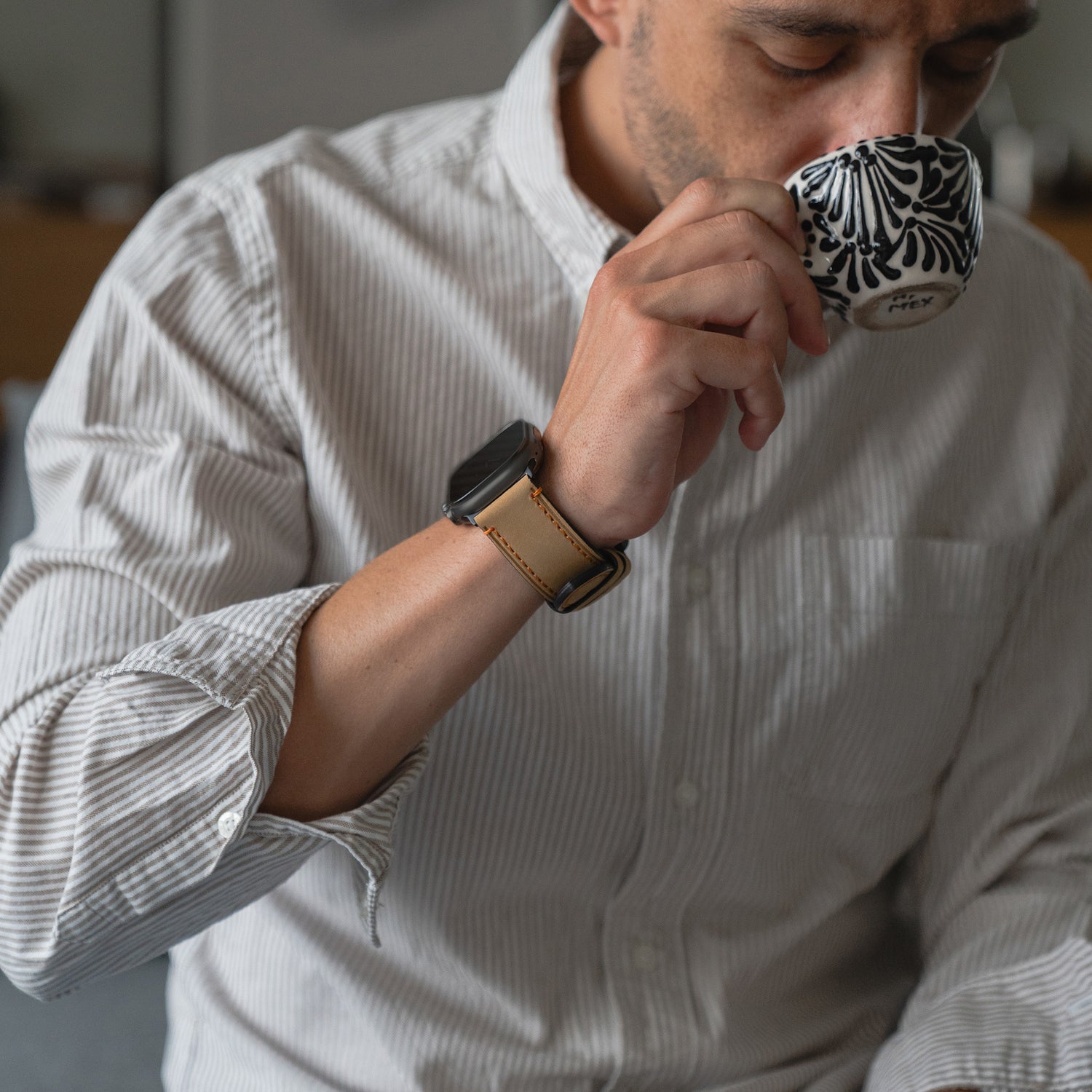 Mann im gestreiften Hemd trinkt aus einem gemusterten Becher, trägt ein Arktisband Apple Watch Ultra PALERMO GRANDE Lederarmband.