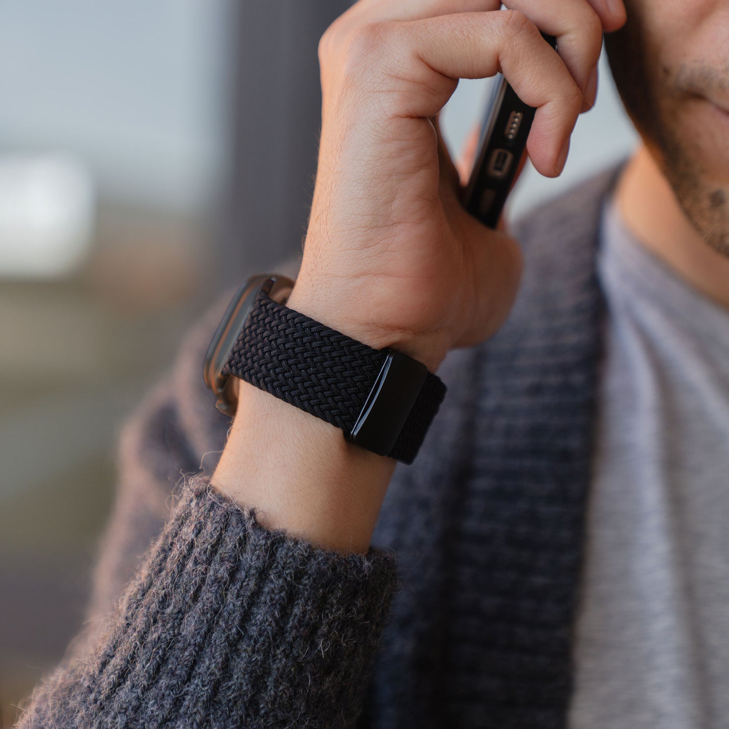 Eine Person, die einen Strickpullover trägt, hält ein Handy ans Ohr; am Handgelenk ist ein „Geflochtenes“ Apple Watch Ultra-Armband „Casual“ von arktisband zu sehen, das sich durch ein stilvolles schwarzes Flechtdesign und einen eleganten Magnetverschluss auszeichnet.