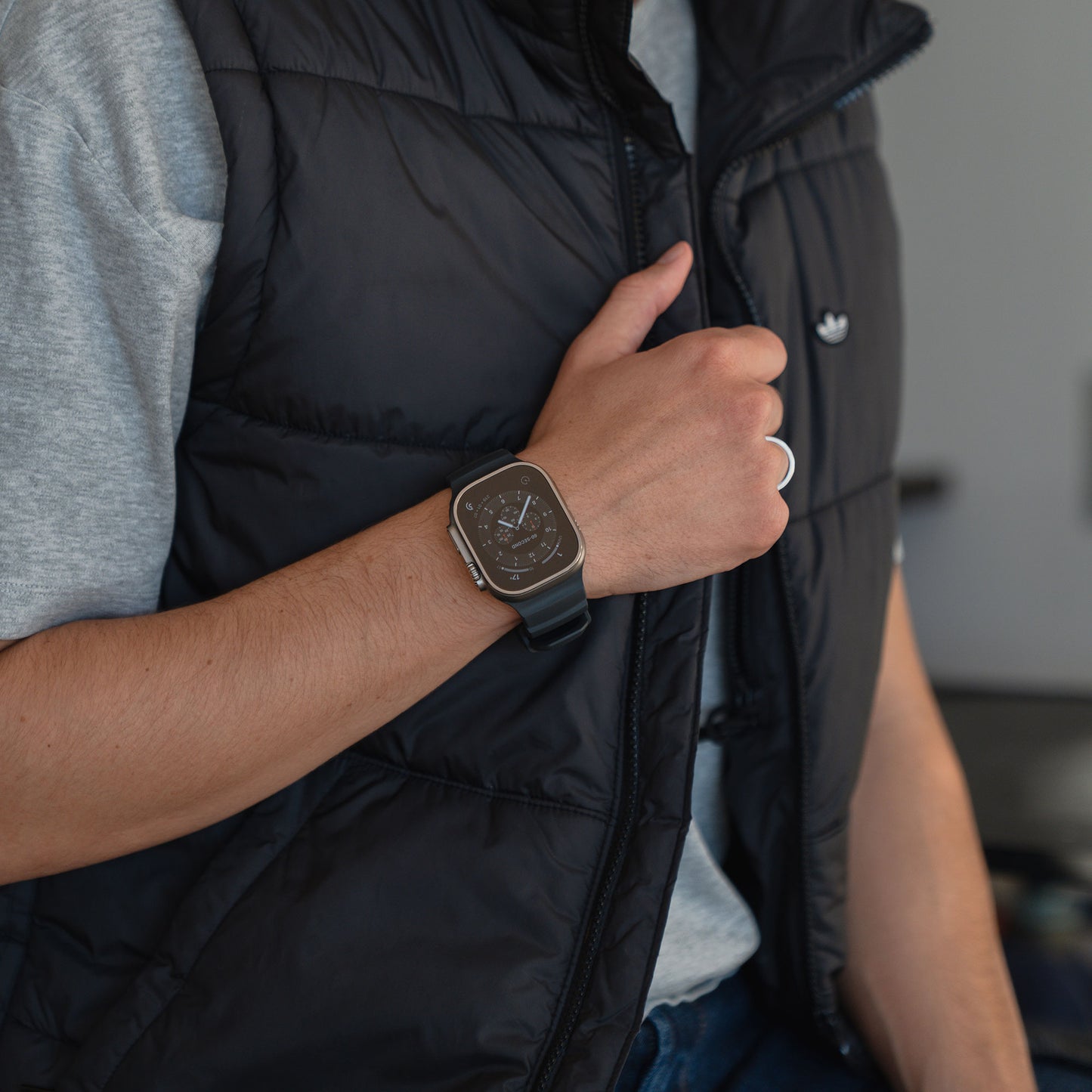 Person, die ein schwarzes arktisband Apple Watch Armband "Flux" von arktisband, ein graues T-Shirt und eine schwarze Weste trägt und die Weste mit einer Hand hält.