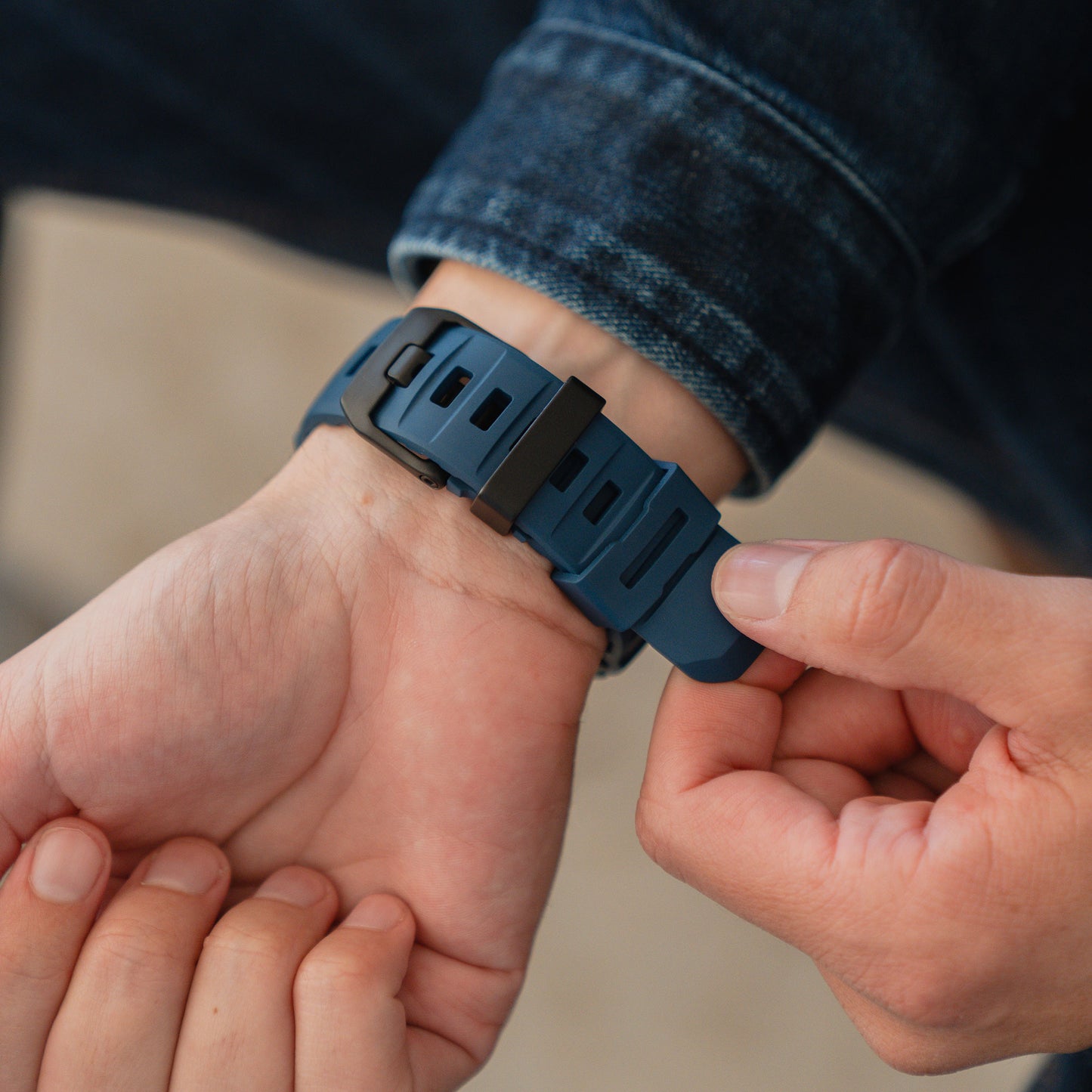 Nahaufnahme von Händen, die ein blaues Arktisband Apple Watch Armband "Flux" mit Edelstahl Schließe an einem Handgelenk einstellen, Person trägt eine Jeansjacke.