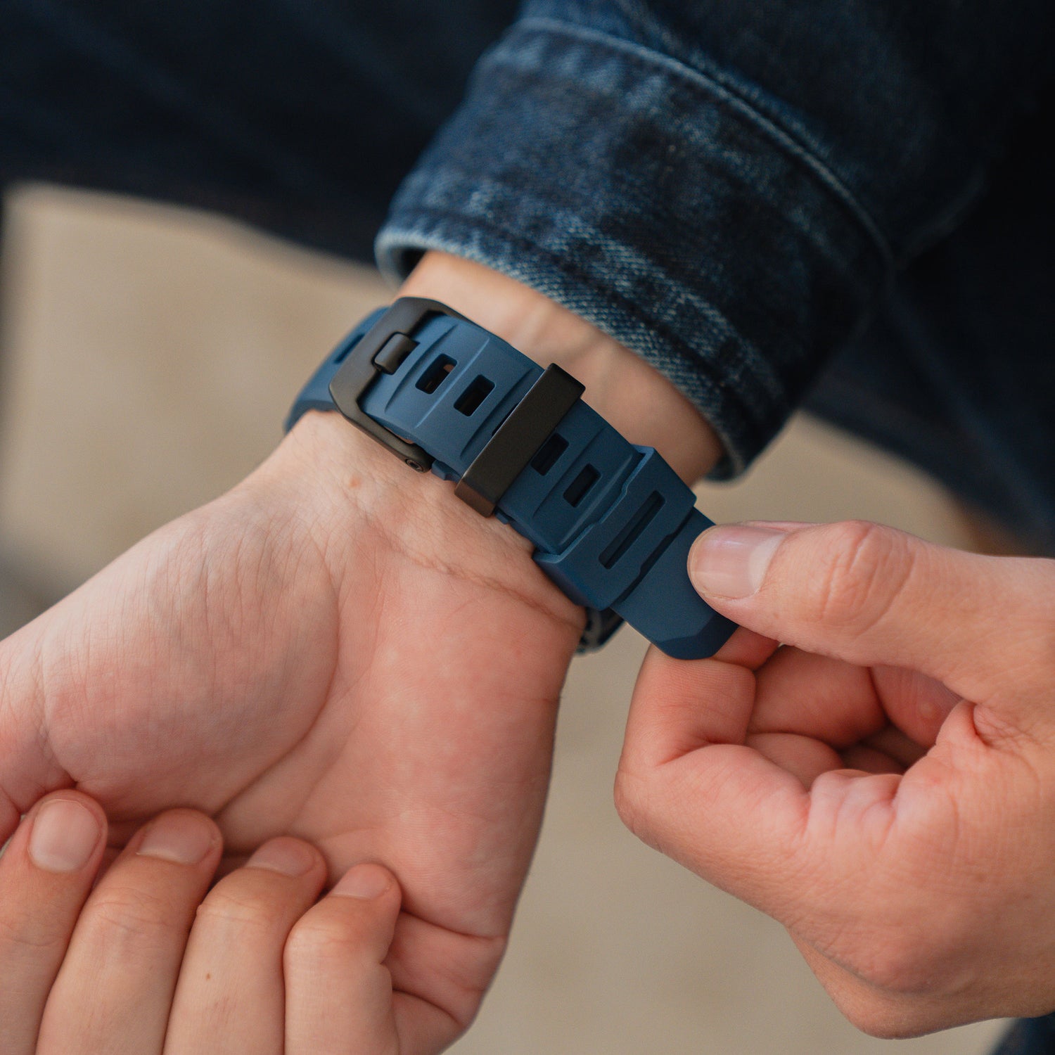 Nahaufnahme von Händen, die ein blaues Arktisband Apple Watch Armband "Flux" mit Edelstahl Schließe an einem Handgelenk einstellen, Person trägt eine Jeansjacke.