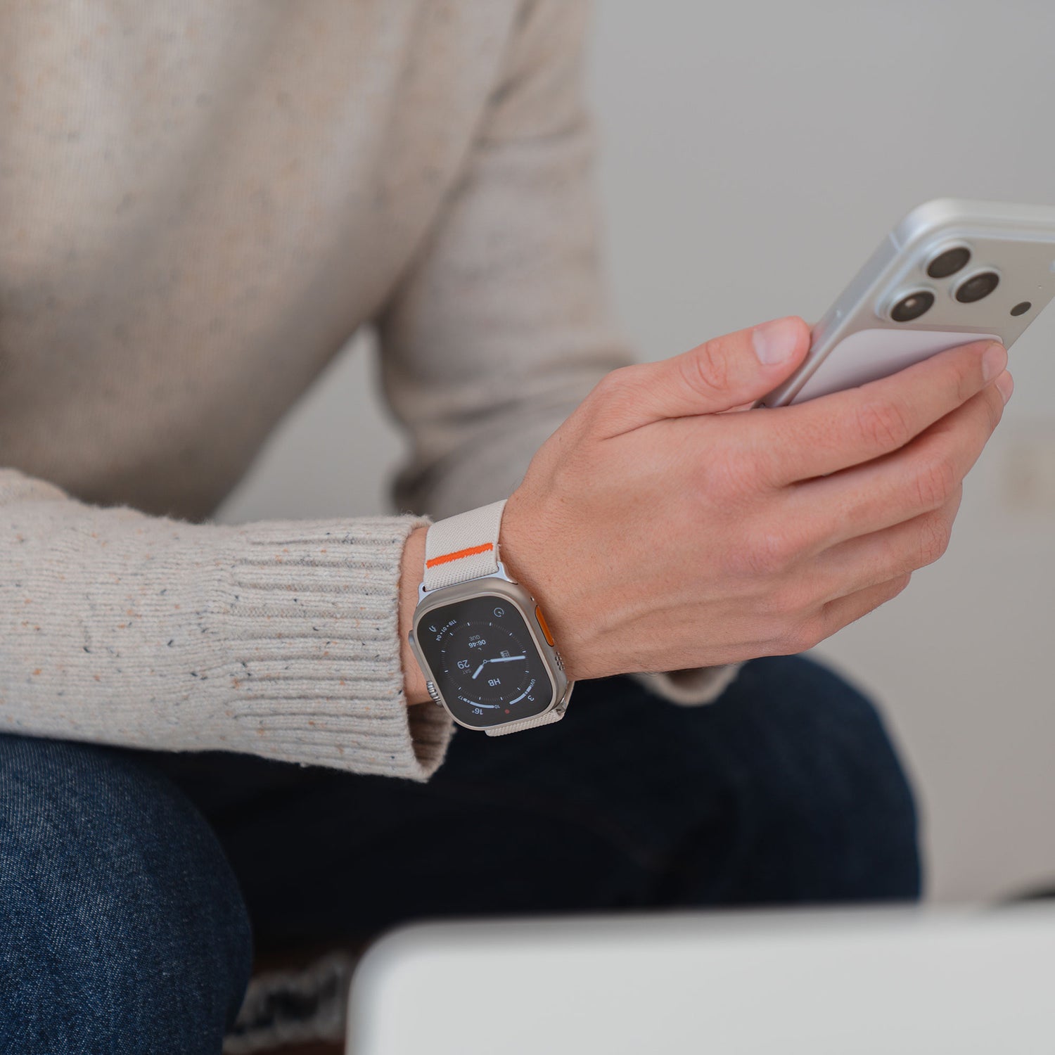 Eine Person, die das Arktisband Apple Watch Nylonarmband "FlexOne" trägt, hält im Sitzen ein Smartphone, bekleidet mit einem beigen Pullover und einer dunklen Jeans.