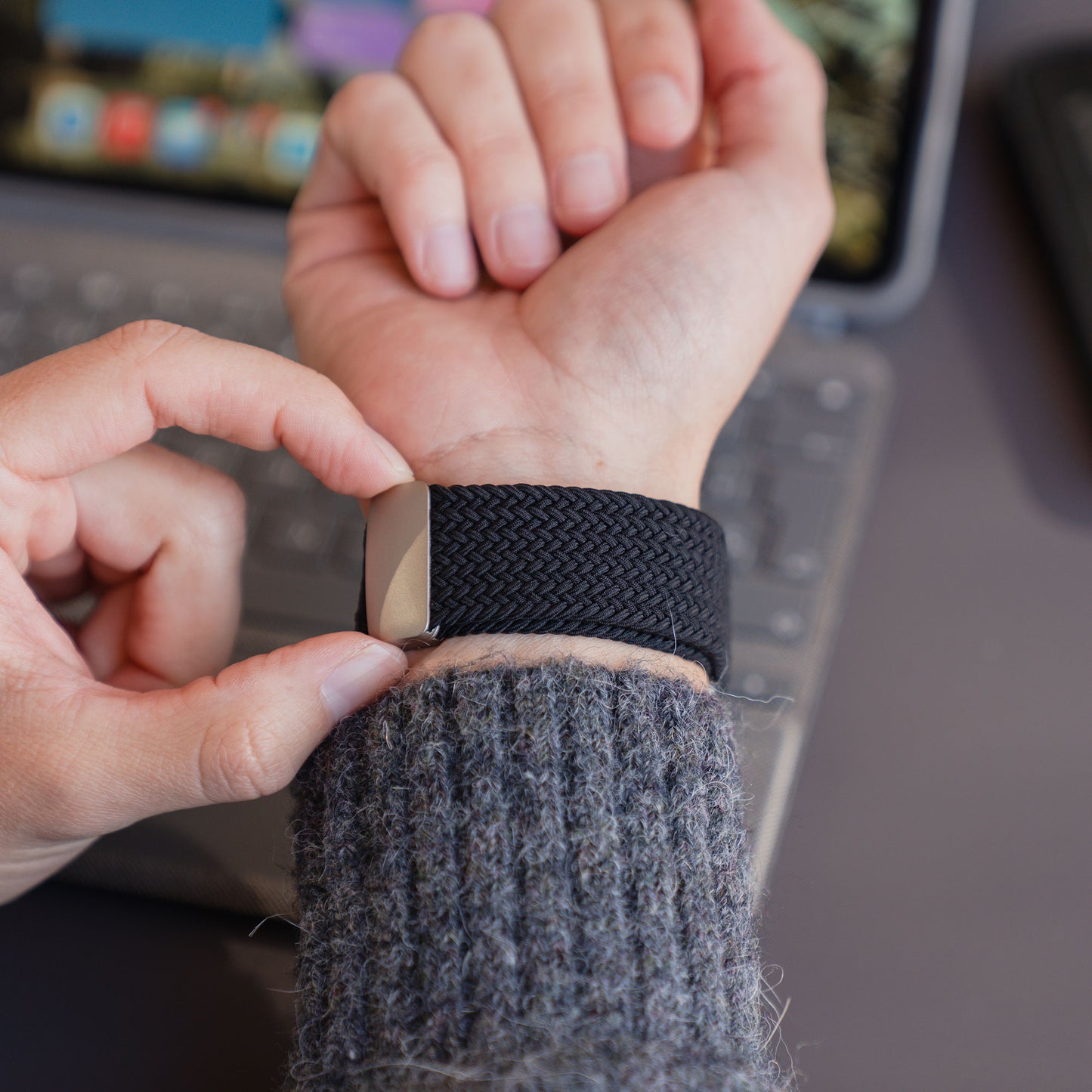 Eine Person sitzt an einem Schreibtisch, im Hintergrund ist ein Laptop zu sehen, und legt ein „Arktisband Geflochtenes Apple Watch Ultra Armband ‚Casual‘“ mit Magnetverschluss an.