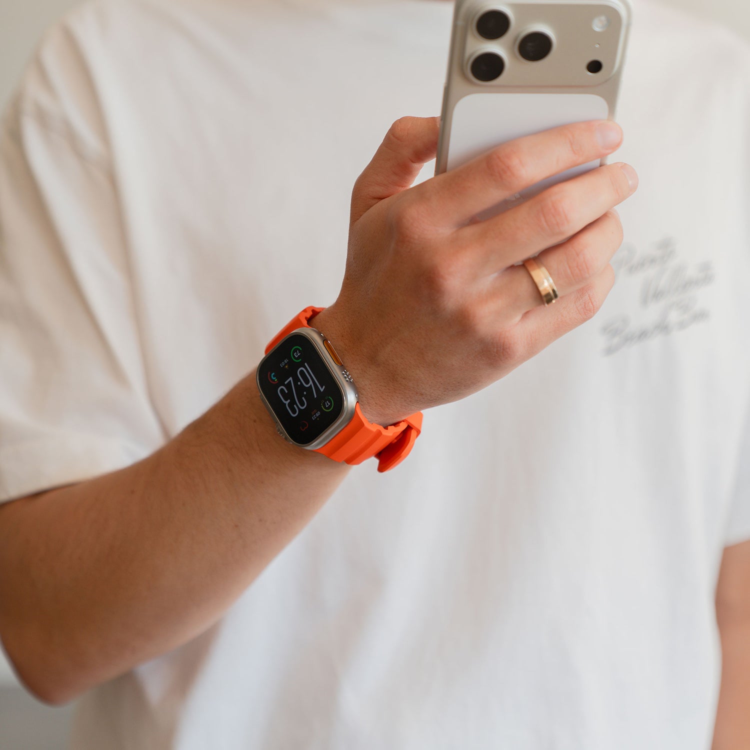 Eine Person in einem weißen T-Shirt trägt das arktisband Apple Watch Armband "Flux" mit orangefarbenem FKM-Armband und Edelstahlverschluss und hält ein Smartphone.