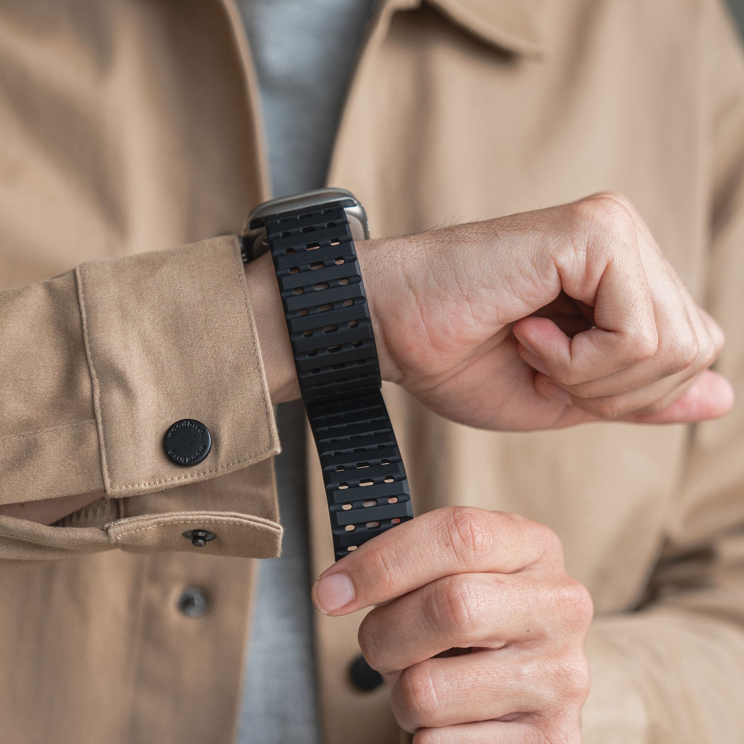 Eine Person trägt eine beige Jacke mit einem schwarzen Knopf an der Manschette und passt ihr magnetisches Silikonarmband „Power“ für die Apple Watch an ihrem linken Handgelenk an, wobei sie den magnetischen Verschluss des Armbands mit der rechten Hand schließt.