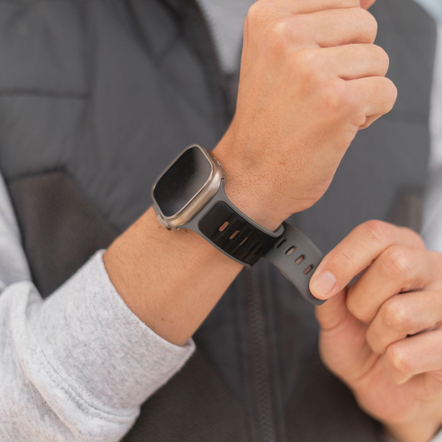 Eine Person in dunkler Weste und hellgrauem Langarmshirt passt das Arktisband Apple Watch Silikonarmband „Swift“, ein schweißabsorbierendes und atmungsaktives Silikonarmband, an ihrem Handgelenk an.