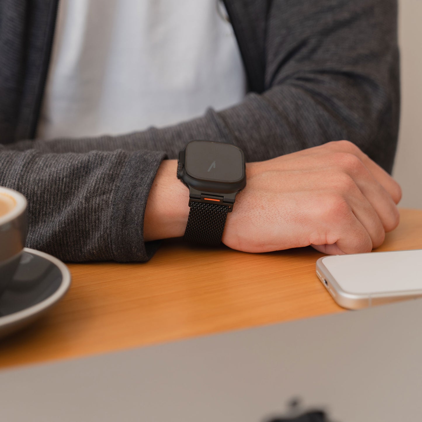 Eine Person, die ein arktisband Apple Watch Milanaise Armband "Bold SE" trägt, sitzt an einem Schreibtisch mit einem Laptop, einem Telefon und einer Tasse Kaffee.