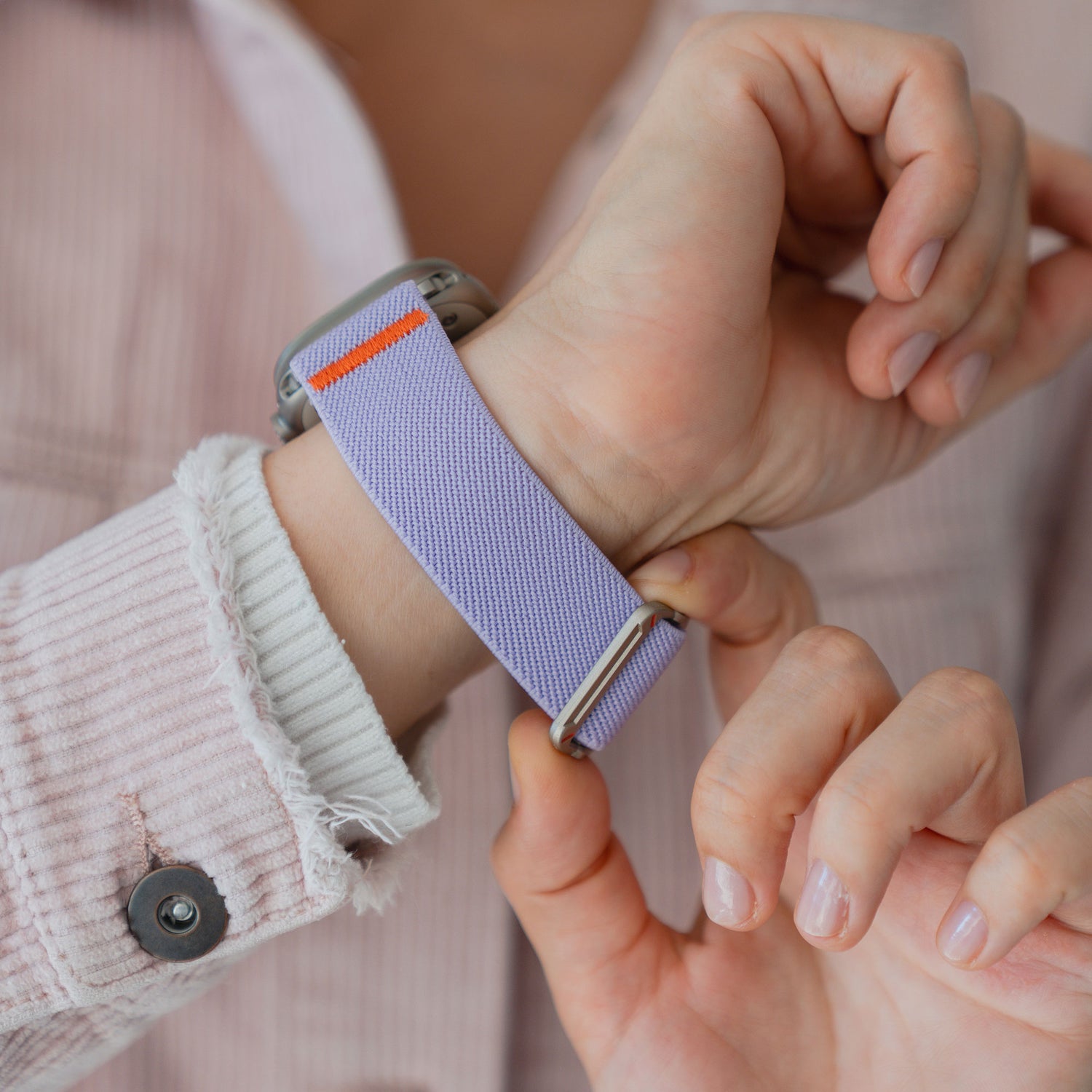 Eine Person befestigt ein Arktisband Apple Watch Nylonarmband "FlexOne" in Lavendel an ihrem Handgelenk, während sie ein hellrosa Cordhemd trägt.