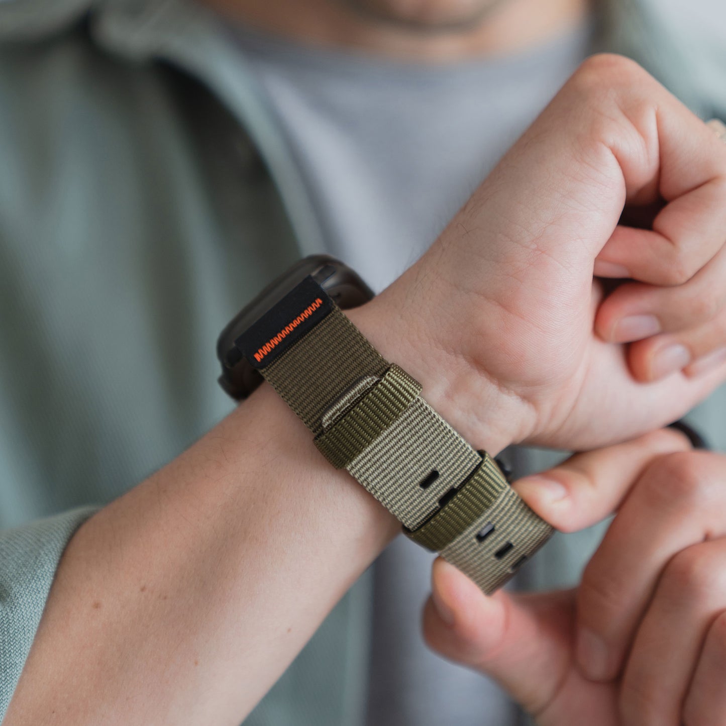 Eine Person befestigt das arktisband Apple Watch Armband "Terra" - ein extrabreites Nylonarmband - an ihrem Handgelenk, während sie ein hellgrünes Hemd trägt und so eine stilvolle Ergänzung für jede Apple Watch Ultra schafft.