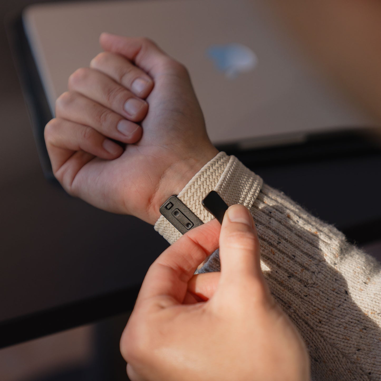 Eine Person legt sich ein „Geflochtenes Apple Watch Ultra Armband ‚Casual‘“ von arktisband in Beige mit Magnetverschluss am Handgelenk an, im Hintergrund ist ein verschwommener Laptop zu sehen.
