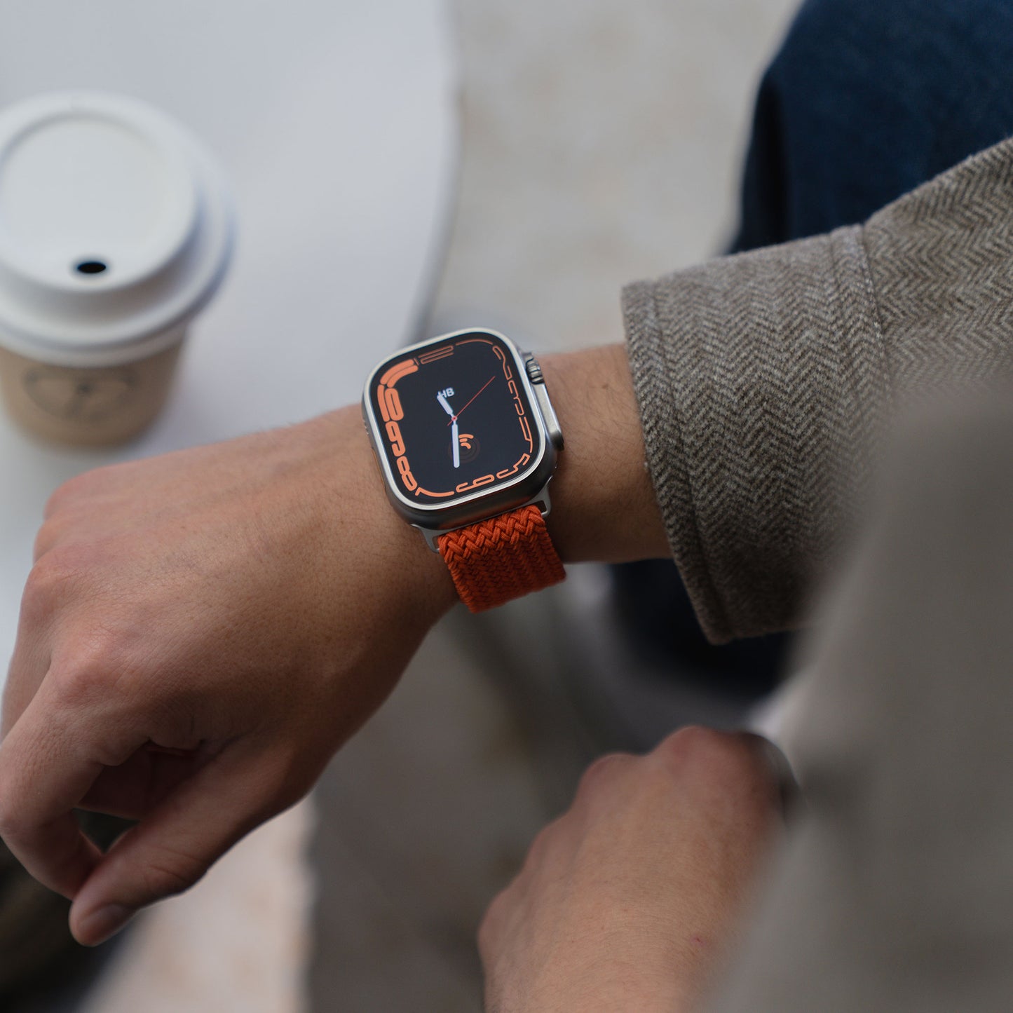 Eine Person, die ein „Geflochtenes“ Arktisband für die Apple Watch Ultra „Casual“ mit Magnetverschluss in Orange trägt und eine Kaffeetasse auf einem weißen Tisch in der Hand hält.