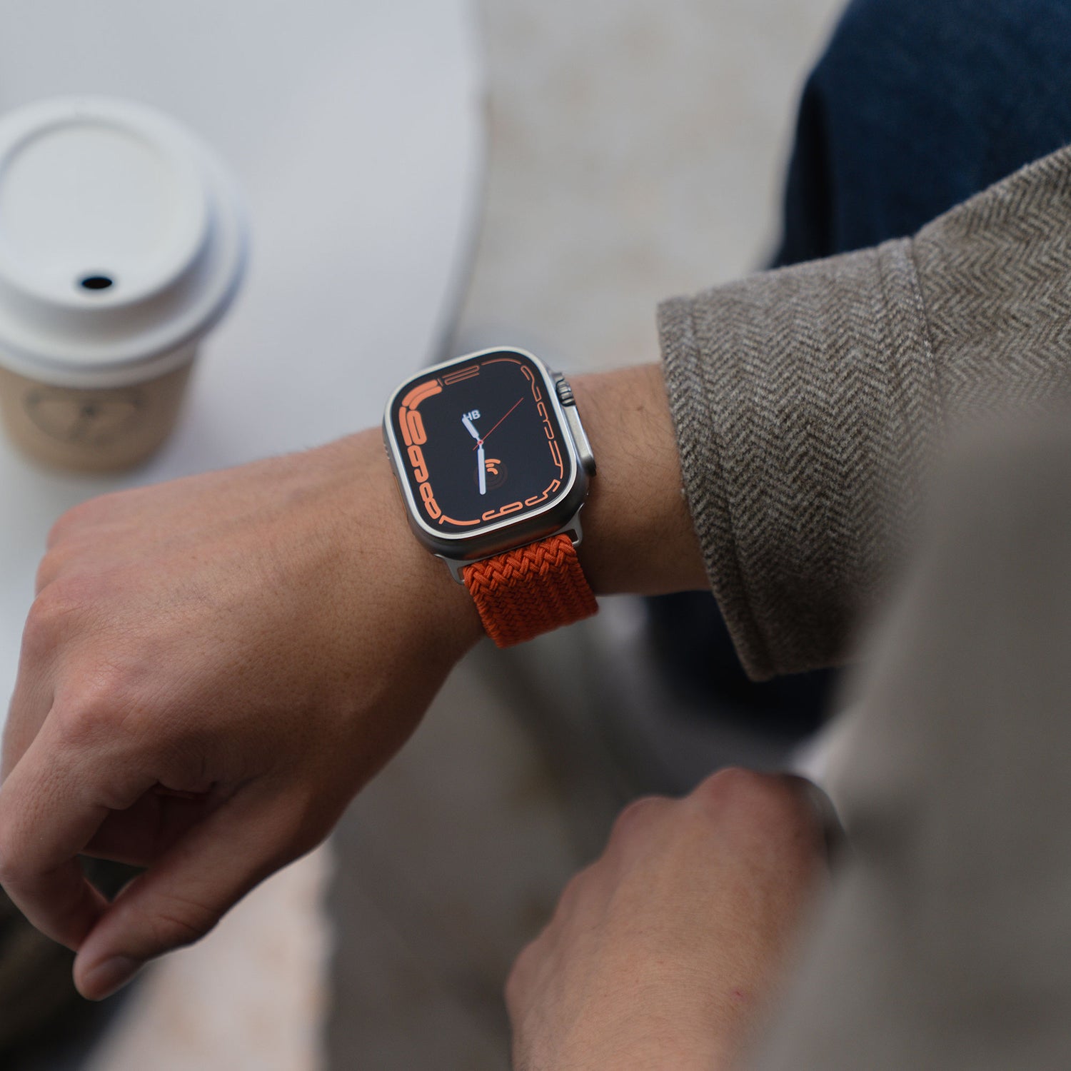 Eine Person, die ein „Geflochtenes“ Arktisband für die Apple Watch Ultra „Casual“ mit Magnetverschluss in Orange trägt und eine Kaffeetasse auf einem weißen Tisch in der Hand hält.