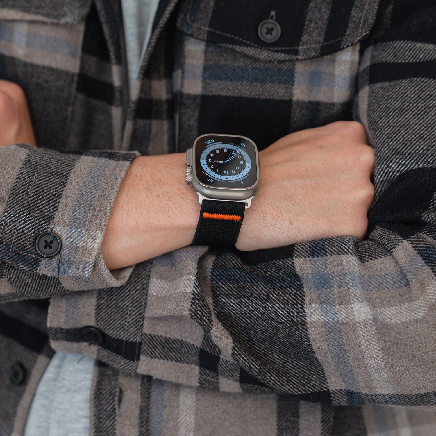Eine Person in einem karierten Hemd verschränkt die Arme und trägt das Arktisband Apple Watch Nylonarmband "FlexOne" mit einem schwarz-orangenen Band.