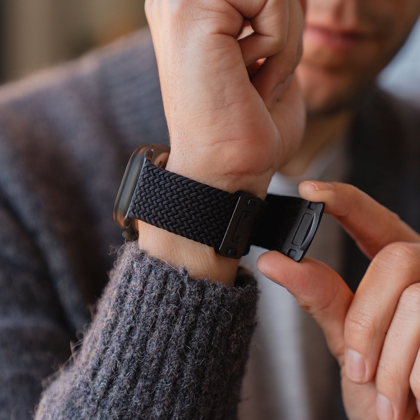 Eine Person legt sich das schwarze Arktisband „Geflochtenes Apple Watch Ultra Armband ‚Casual‘“ mit Magnetverschluss am Handgelenk an, während sie einen grauen Strickpullover trägt.