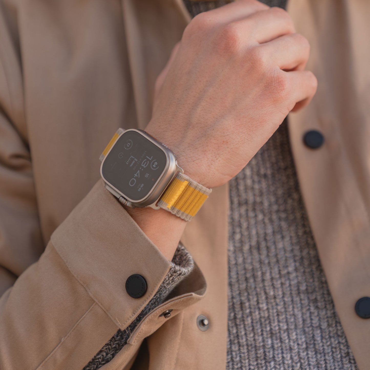 Nahaufnahme einer Person, die das arktisband Apple Watch Armband "CoreX" im stylischen Gold- und Silber-Wave-Look trägt, gepaart mit einem hellbraunen Mantel und einem grauen Pullover.