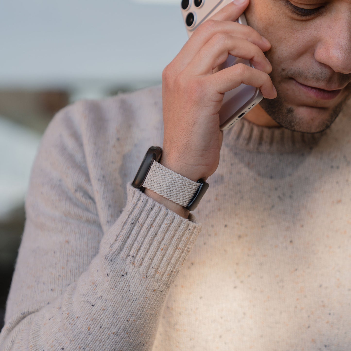 Eine Person, die einen beigen Pullover und ein Arktisband „Geflochtenes Apple Watch Ultra Armband ‚Casual‘“ mit Magnetverschluss trägt, telefoniert mit einem Smartphone, wobei nur der untere Teil ihres Gesichts zu sehen ist.