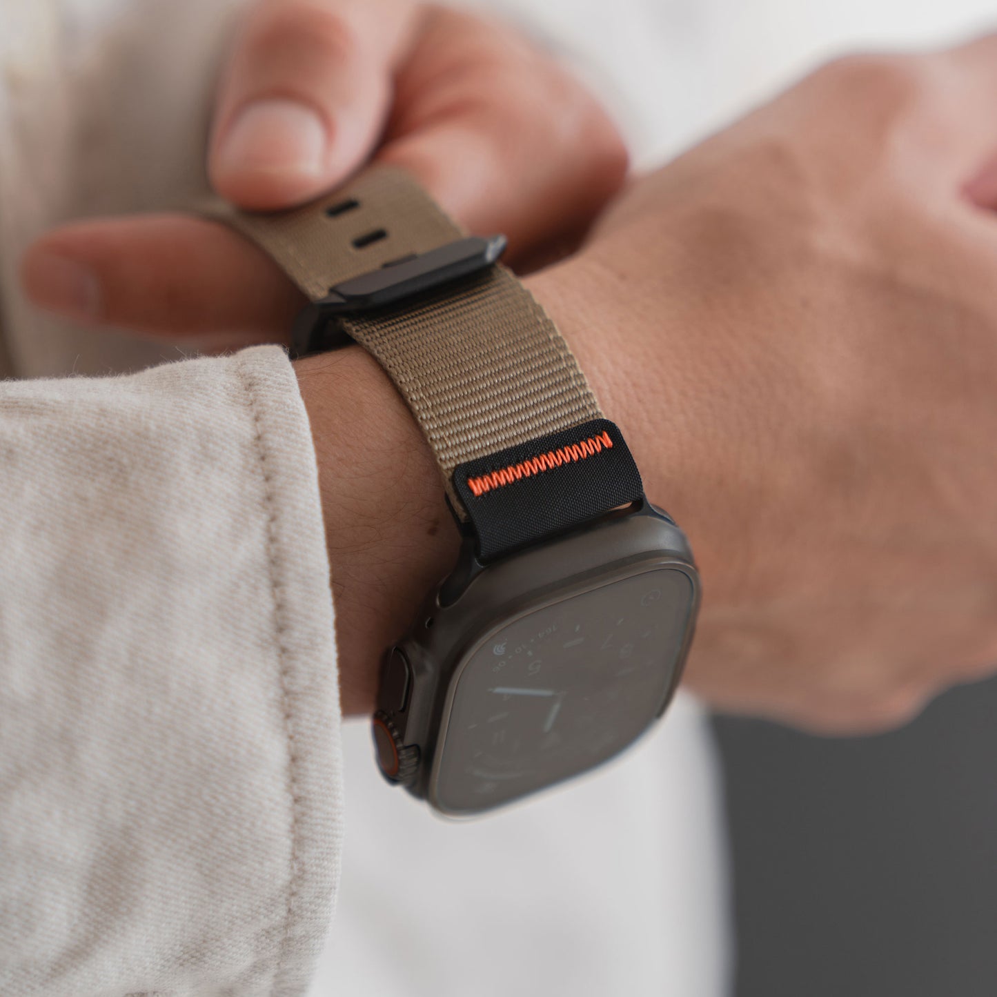Jemand passt sein arktisband Apple Watch Armband "Terra" mit beigem Outdoor-Nylongewebe, extrabreitem 24-mm-Gewebe und orangefarbenen Nähten an sein Handgelenk an.