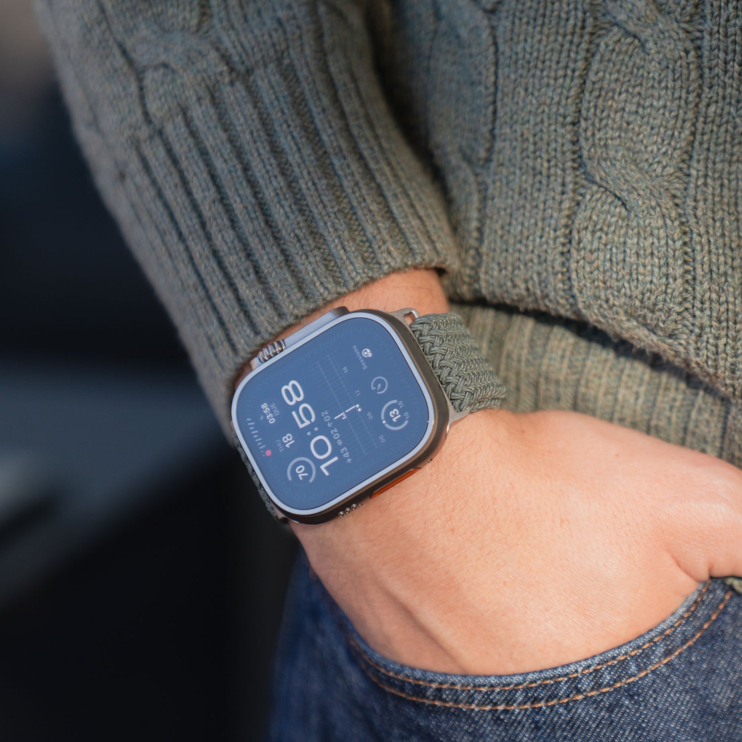 Eine Person, die eine graue Apple Watch mit dem „Geflochtenes Apple Watch Ultra Armband ‚Casual‘ mit Magnetverschluss“ von arktisband trägt, sowie einen grünen Strickpullover, wobei eine Hand in der Tasche einer blauen Jeans steckt.