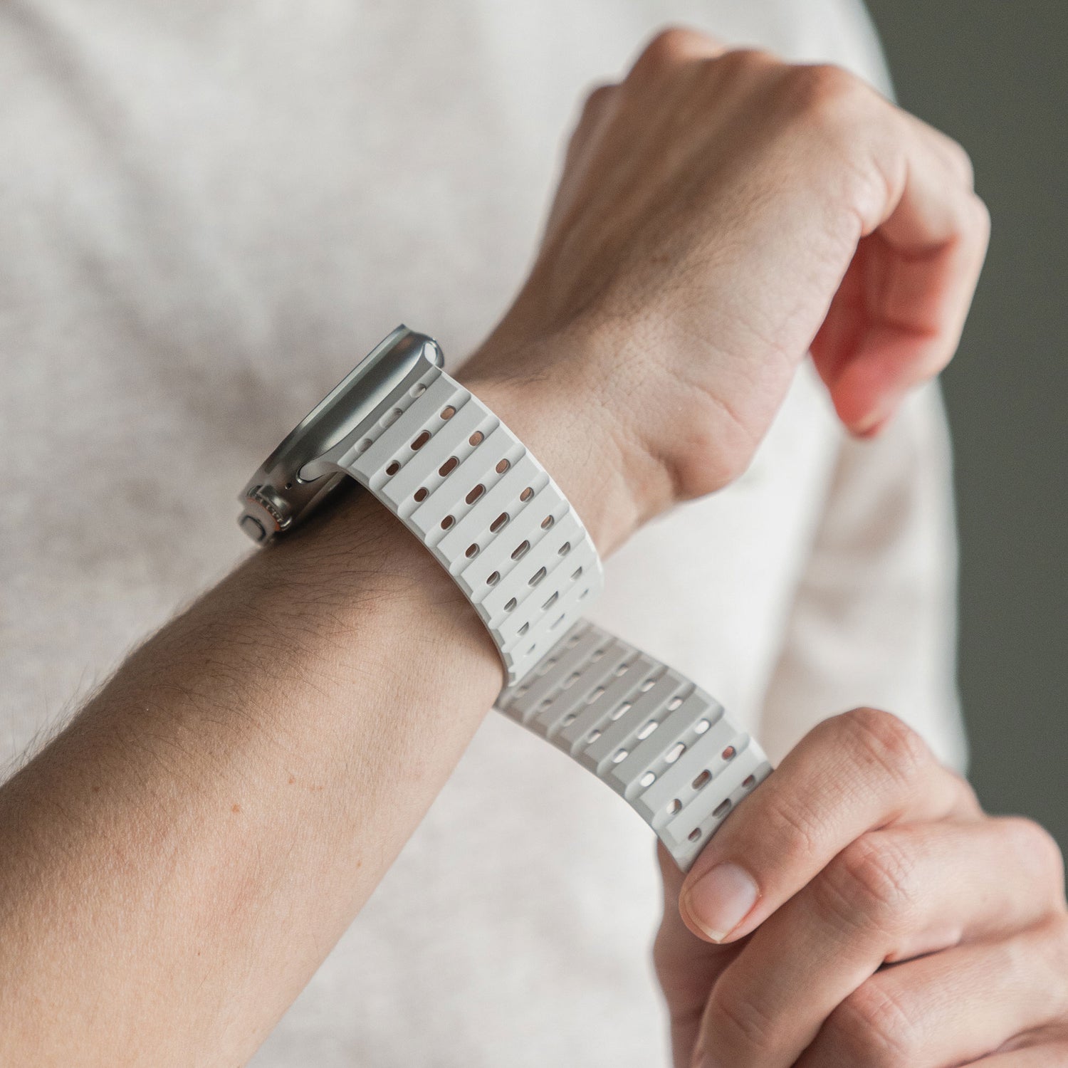 Eine Person befestigt ein arktisband Apple Watch magnetisches Silikonarmband „Power“ von arktis am linken Handgelenk. Das weiße perforierte Band wird mit einem hellen Hemd getragen, wobei der Hintergrund unscharf erscheint.