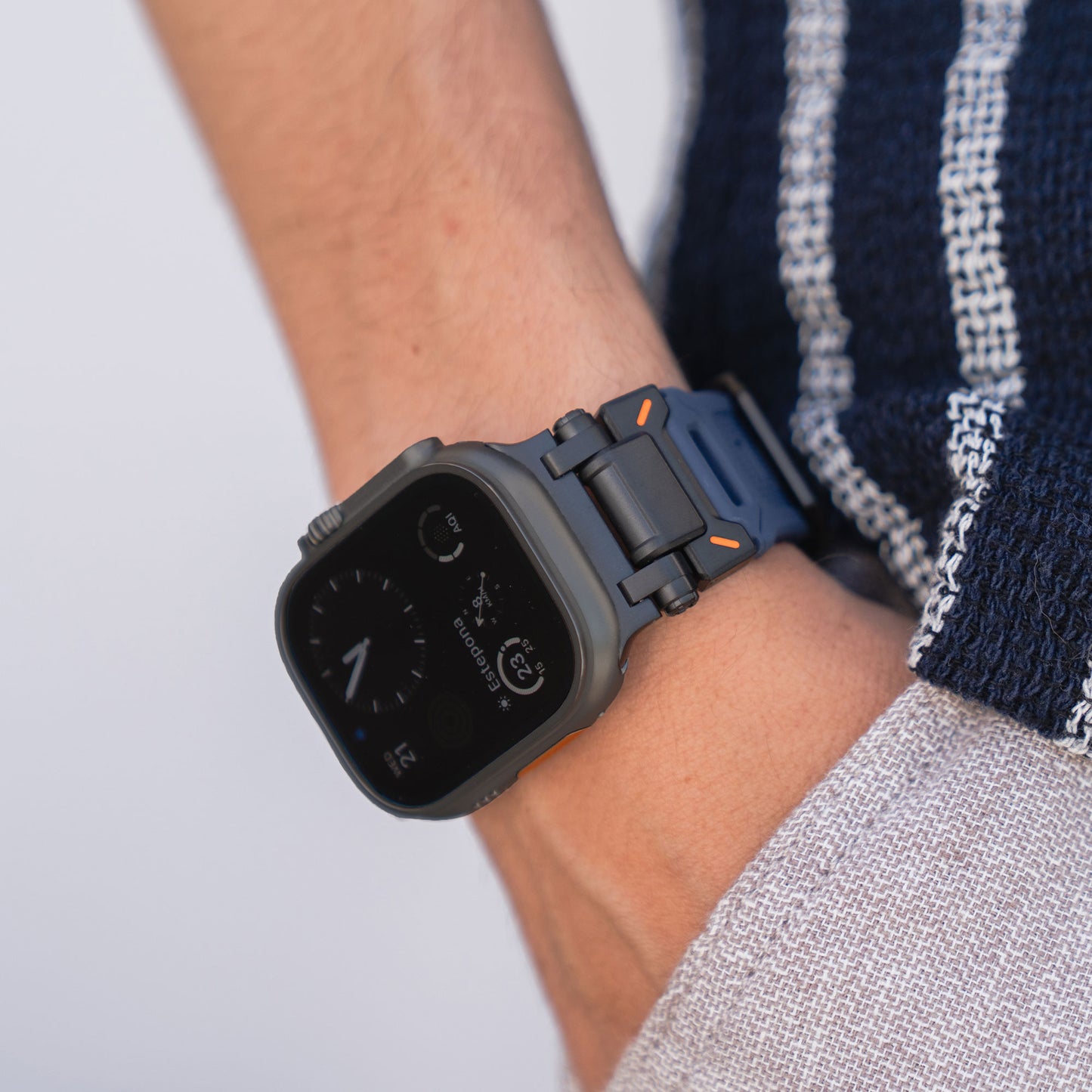 Eine Person trägt ein Arktisband Apple Watch Ultra Armband „Atlas“ mit orangefarbenen Akzenten am linken Handgelenk, dazu ein marineblau-weiß gestreiftes Hemd und eine hellgraue Hose. Das Zifferblatt zeigt die Uhrzeit und digitale Details an.