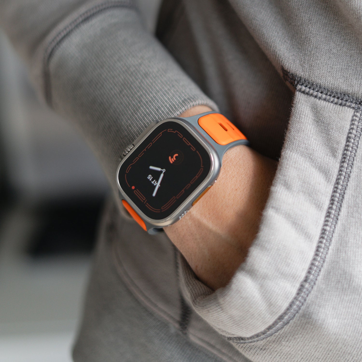 Eine Person in einem grauen Sweatshirt, die Hand in der Tasche, zeigt ein arktisband Apple Watch Silikonarmband „Swift“ – ein orangefarbenes, schweißabsorbierendes und atmungsaktives Silikonarmband – mit einem dunklen Bildschirm, auf dem Fitnessdaten angezeigt werden.