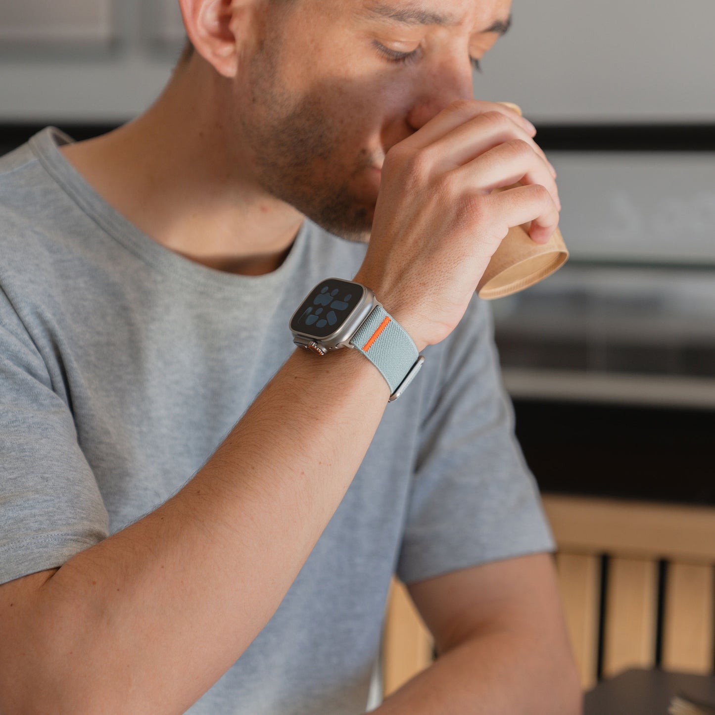 Mann im grauen Hemd trinkt aus einem Pappbecher, trägt eine Apple Watch Nylonarmband "FlexOne" mit einem hellblauen Armband.
