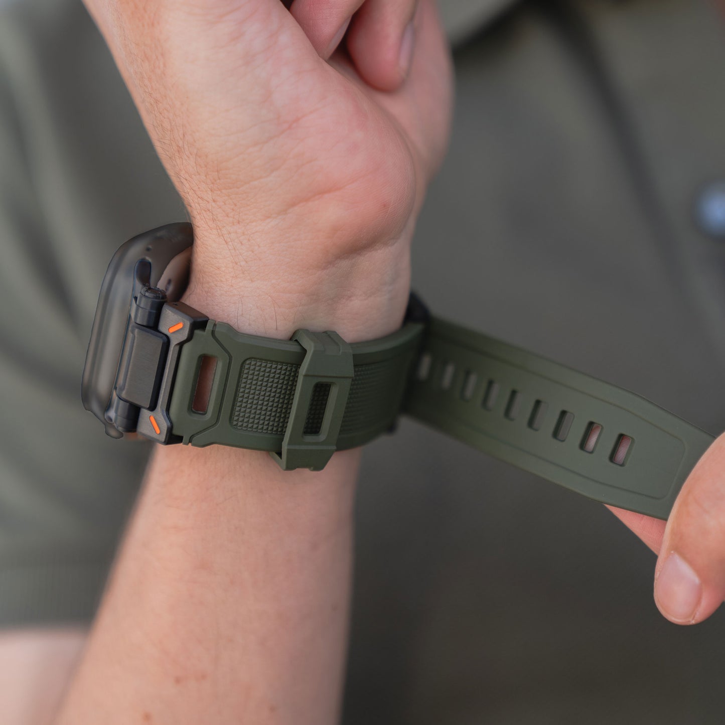 Eine Person befestigt ein Arktisband Apple Watch Ultra Armband „Atlas“ mit robustem, grünem Band an ihrem Handgelenk. Der Fokus liegt auf dem Armband und dem Verschluss, vor dem unscharfen Hintergrund ist ein Ausschnitt eines grünen Hemdes zu sehen.