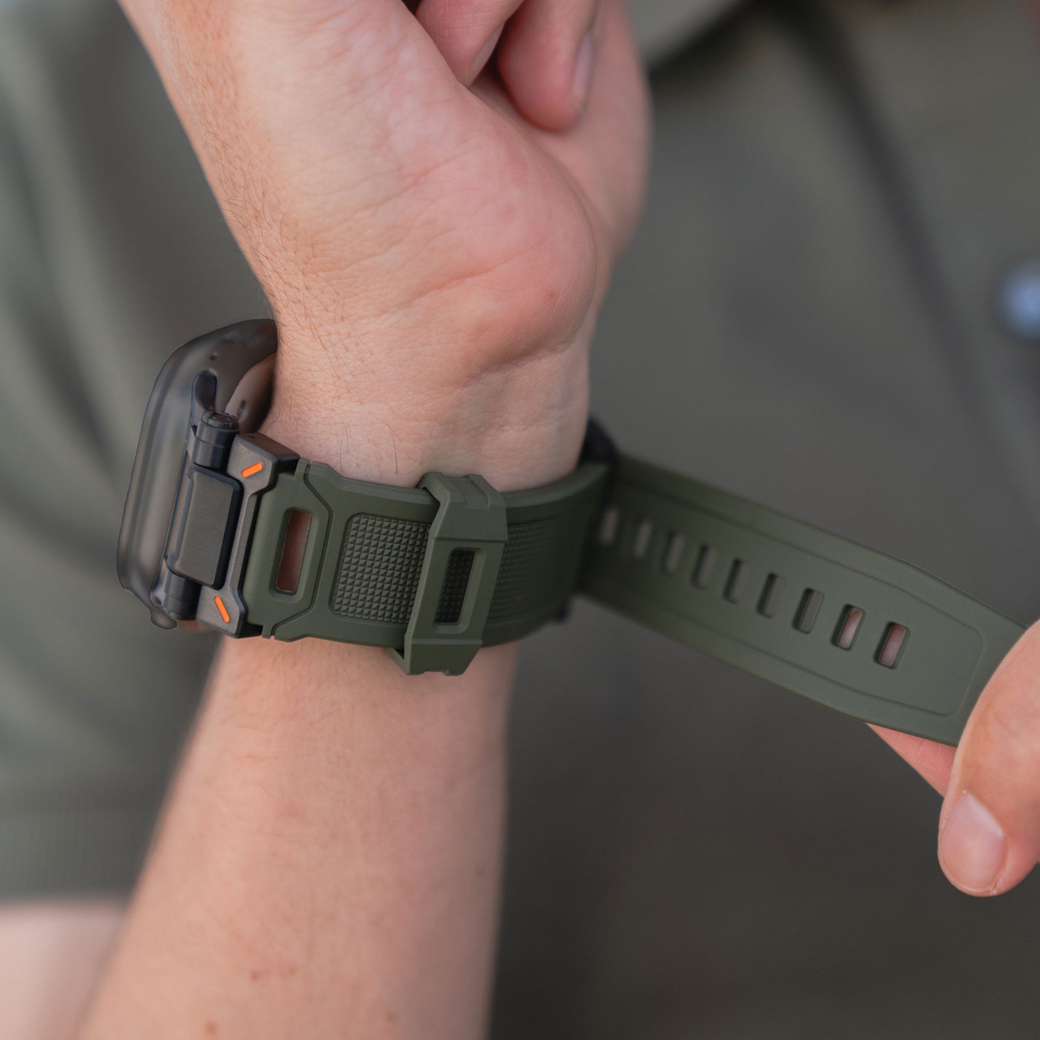 Eine Person befestigt ein Arktisband Apple Watch Ultra Armband „Atlas“ mit robustem, grünem Band an ihrem Handgelenk. Der Fokus liegt auf dem Armband und dem Verschluss, vor dem unscharfen Hintergrund ist ein Ausschnitt eines grünen Hemdes zu sehen.