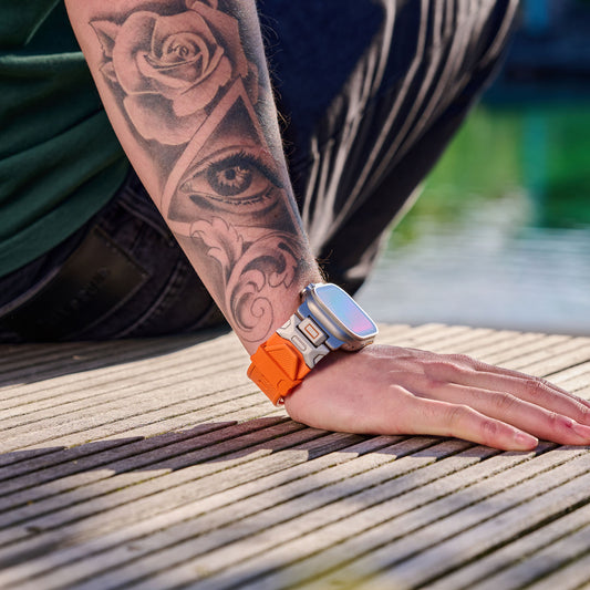 Eine Person mit tätowiertem Arm ruht auf einem Holzbrett am Wasser. Sie trägt ein Arktisband Apple Watch Ultra Armband „Alpha“ mit orangefarbenem FKM-Kunststoffband. Ein Teil ihres grünen Hemdes ist sichtbar.