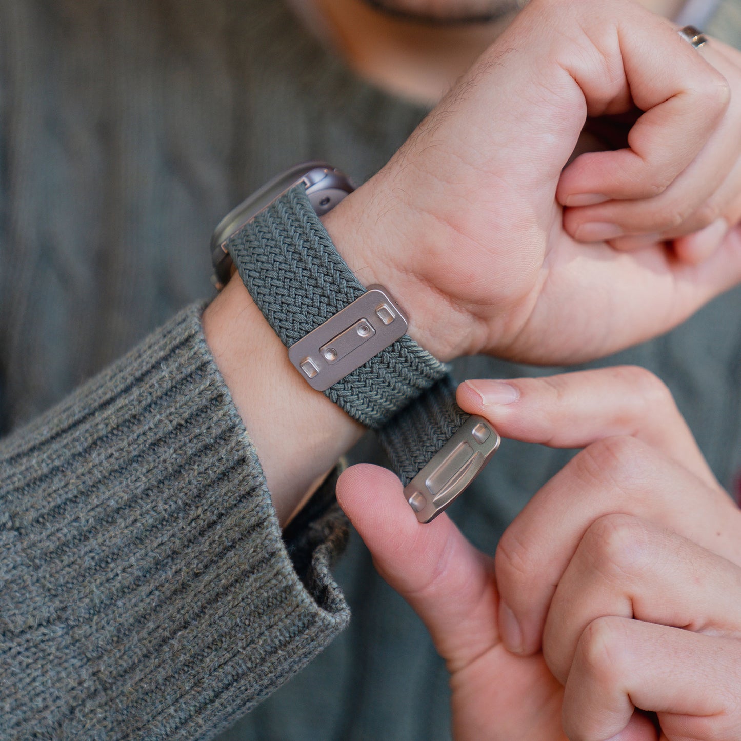 Nahaufnahme einer Person, die das Arktisband „Geflochtenes Apple Watch Ultra-Armband ‚Casual‘“ mit Magnetverschluss in Grau am Handgelenk befestigt, während sie einen Strickpullover trägt, der perfekt zu ihrem Apple Watch Ultra-Armband passt.