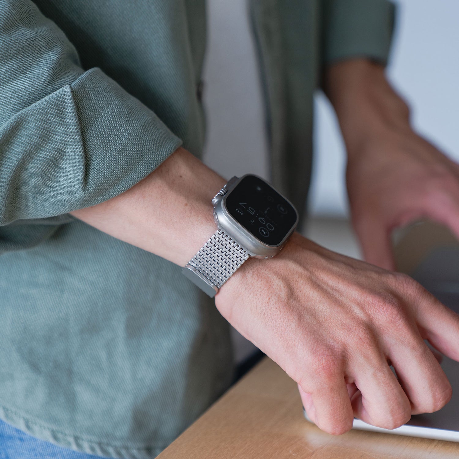 Eine Person, die das arktisband Apple Watch Edelstahlarmband „Aurora“ von arktisband trägt, sitzt an einem Schreibtisch mit einem Laptop und trägt eine hellgrüne Jacke und ein weißes Hemd.