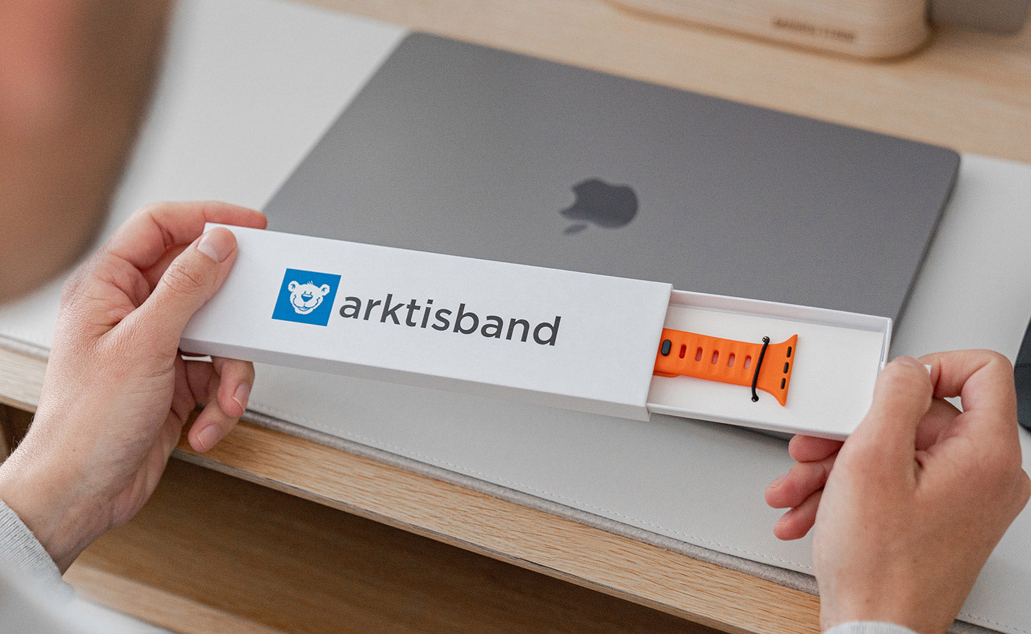 Person hält eine weiße Arktisband-Box mit einem orangefarbenen Uhrenarmband neben einem geschlossenen MacBook.