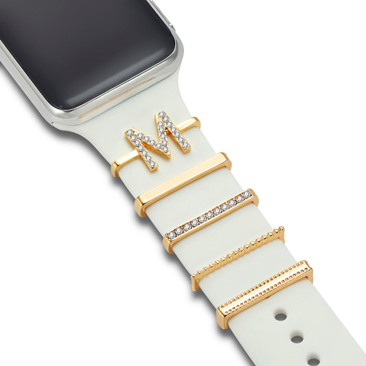 Ein weißes Smartwatch-Armband, verziert mit vier arktisband Apple Watch Charms „M Style“ von arktisband, mit einem Strassbuchstaben M und drei rechteckigen goldenen Bändern mit Edelstahl-, Glas- oder Perlendesigns.