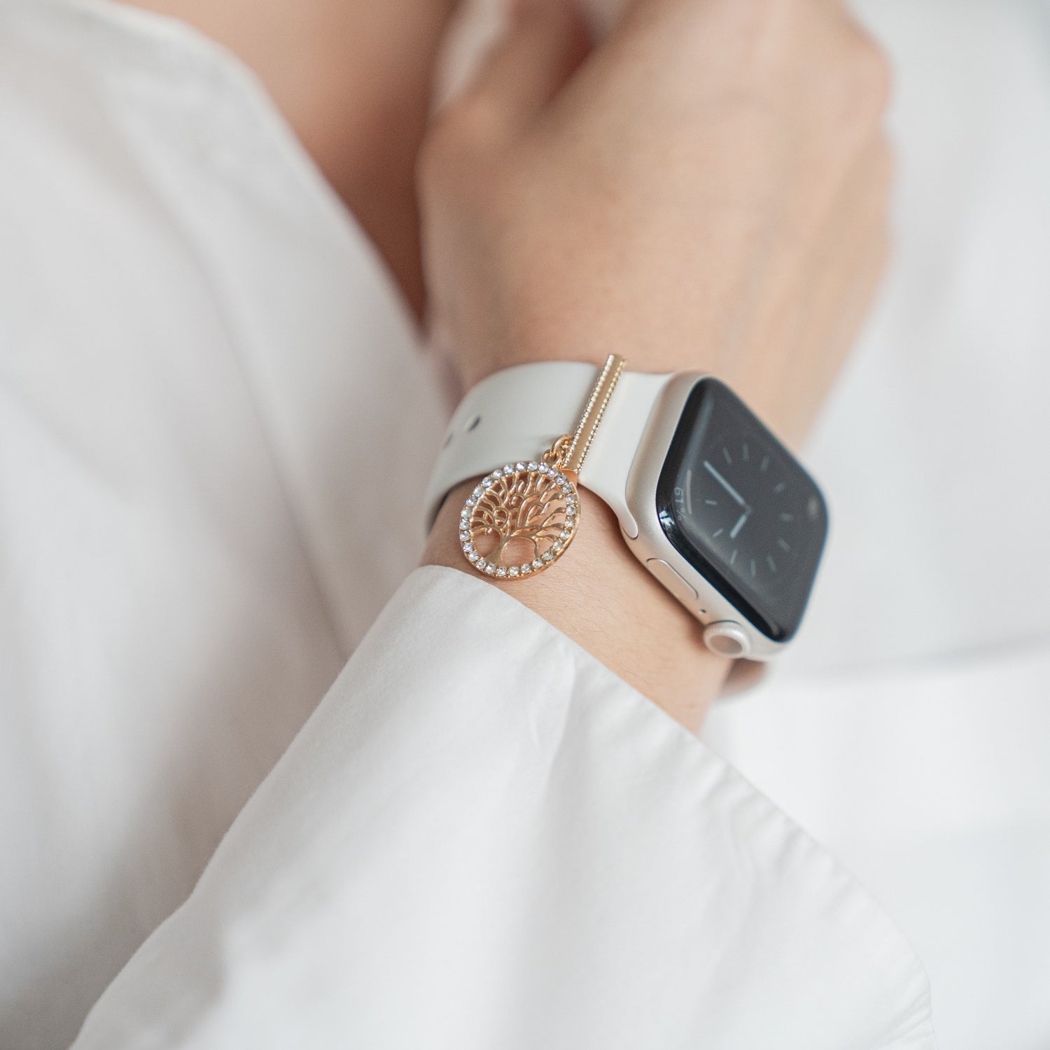 Eine Person trägt eine weiße Smartwatch mit Arktisband Apple Watch Charms „Baum des Lebens“ in Gold über einem weißen Langarmshirt.