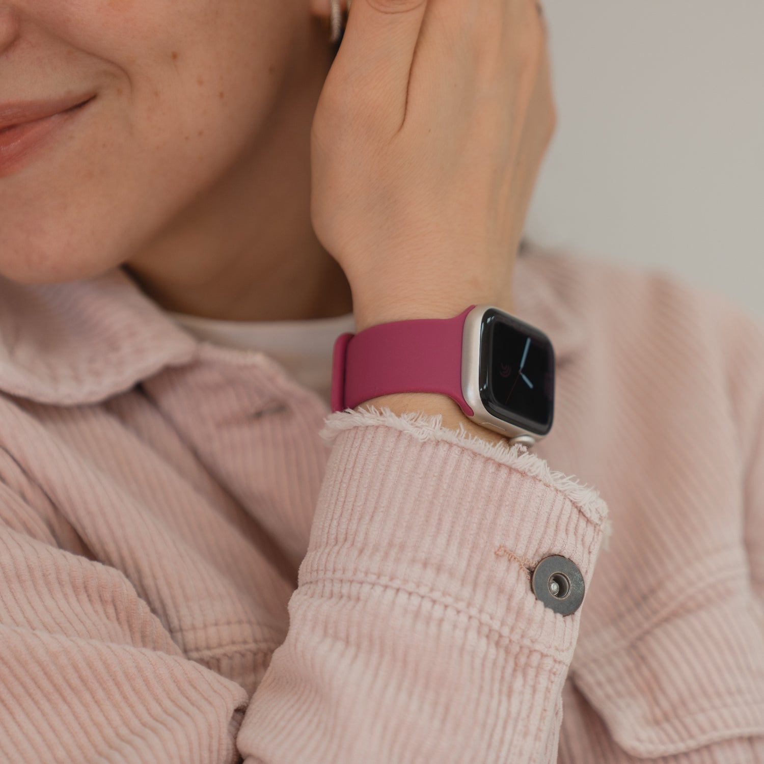 Eine Person in einer rosa Jacke lächelt und berührt ihr Ohr, sie trägt das Arktisband Apple Watch Silikonarmband "Sport" - ideal für sportliche Aktivitäten.