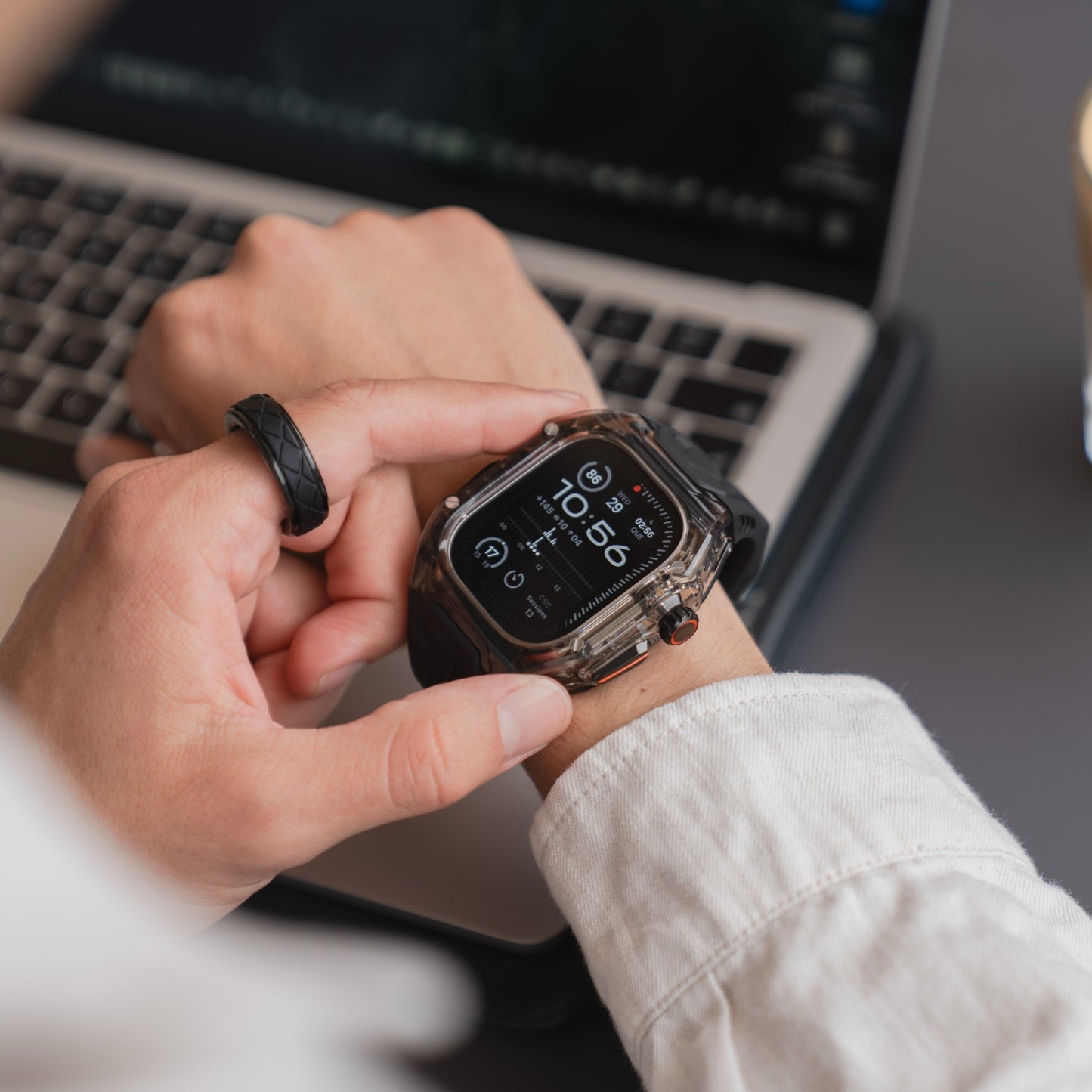 Eine Person überprüft ihre Smartwatch mit dem arktisband Apple Watch Armband "Armor Fusion" an ihrem Handgelenk, während ihr Gerät durch ein transparentes Hardcase an einem modernen Schreibtisch vor einem offenen Laptop geschützt ist.