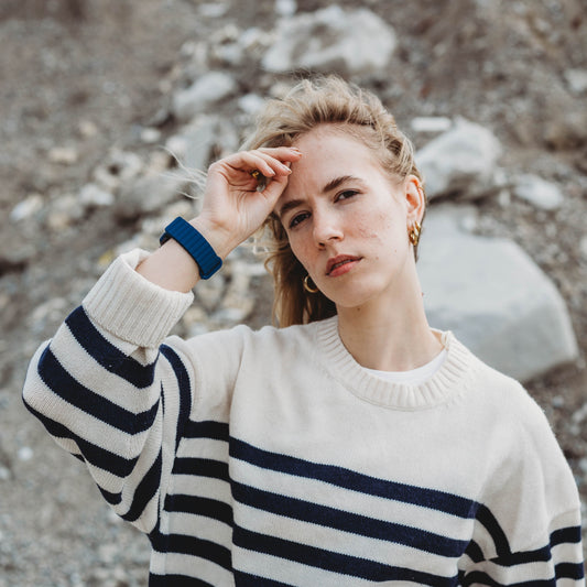 Eine Person mit hellem Haar trägt einen gestreiften Pullover und das arktisband Apple Watch Magnetarmband „Neo“ von arktisband, steht im Freien vor einer felsigen Landschaft, blickt in die Kamera und berührt mit einer Hand ihre Stirn.