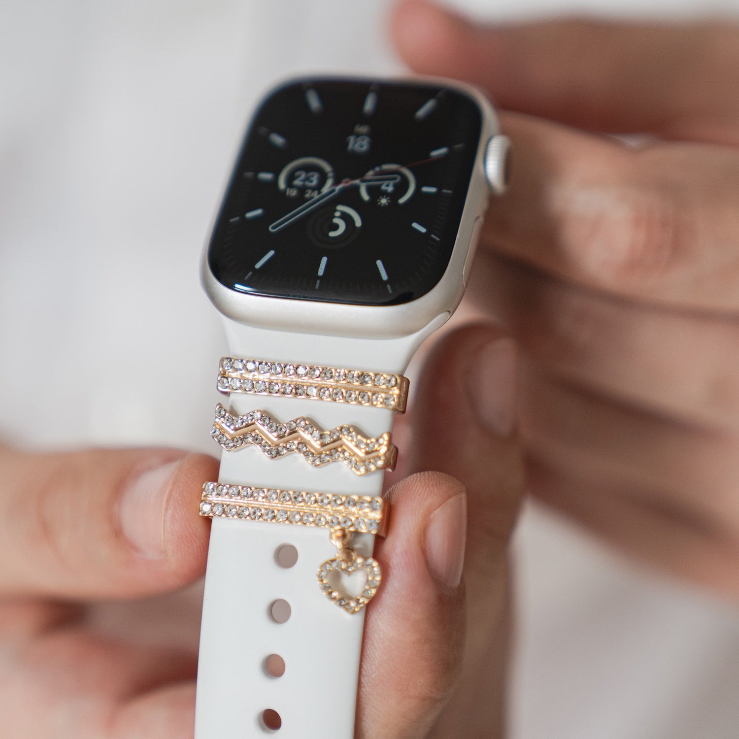 Eine Hand hält die Arktisband-Apple-Watch-Charms „Hanging Heart“ in Gold auf einem weißen Arktisband mit juwelenbesetzten Charms und einem Herz.