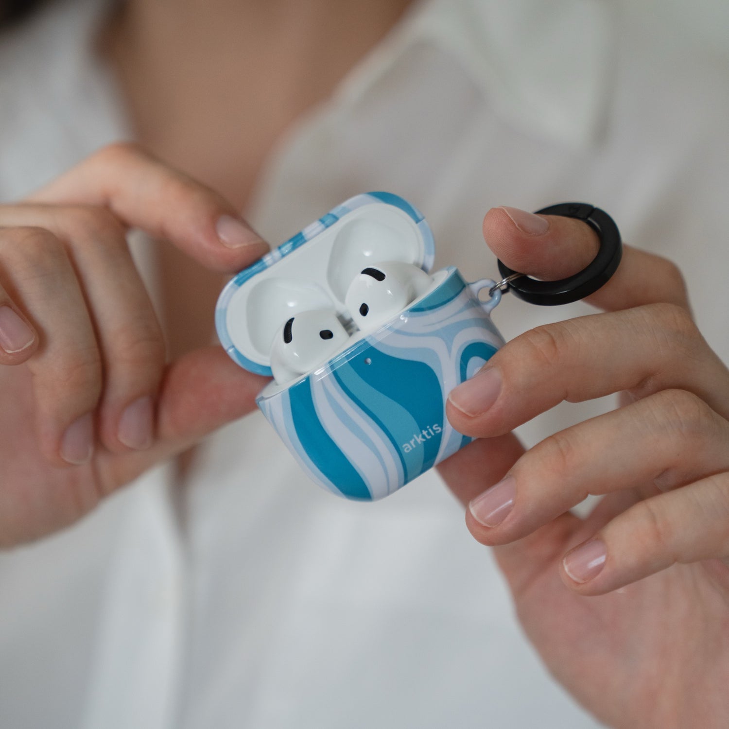 Hände, die eine geöffnete Arktis AirPods 5-Hülle – Blue Lagoon – mit einem blau-weißen Wellenmuster halten, in der sich die Ohrhörer befinden.