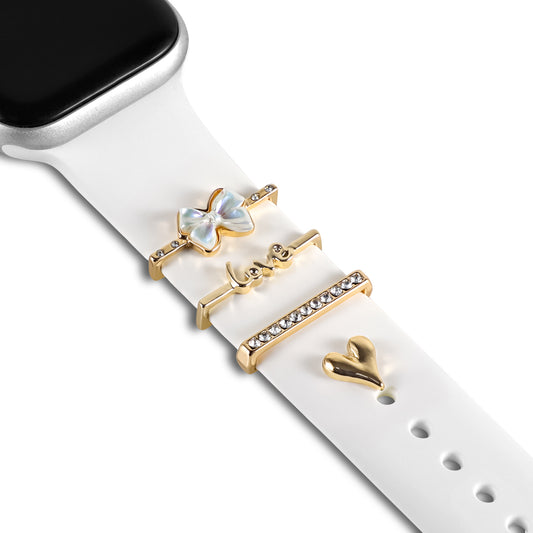 Das arktisband Apple Watch Charms „Love Bow“ Set von arktisband besteht aus einem weißen Armband mit vier goldenen Charms: Schmetterling, „love“-Bar, Kristallleiste und Herz. Die typische Künstlerband-Uhranzeige ist teilweise sichtbar.