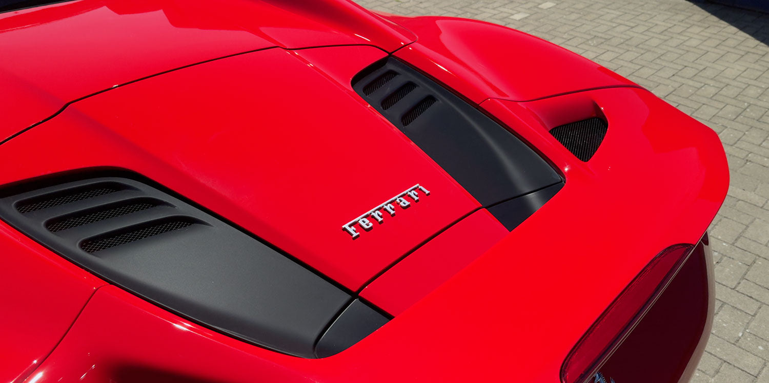 Nahaufnahme der Motorhaube eines roten Ferrari-Sportwagens mit schwarzen Lüftungsschlitzen und dem Ferrari-Logo in der Mitte.