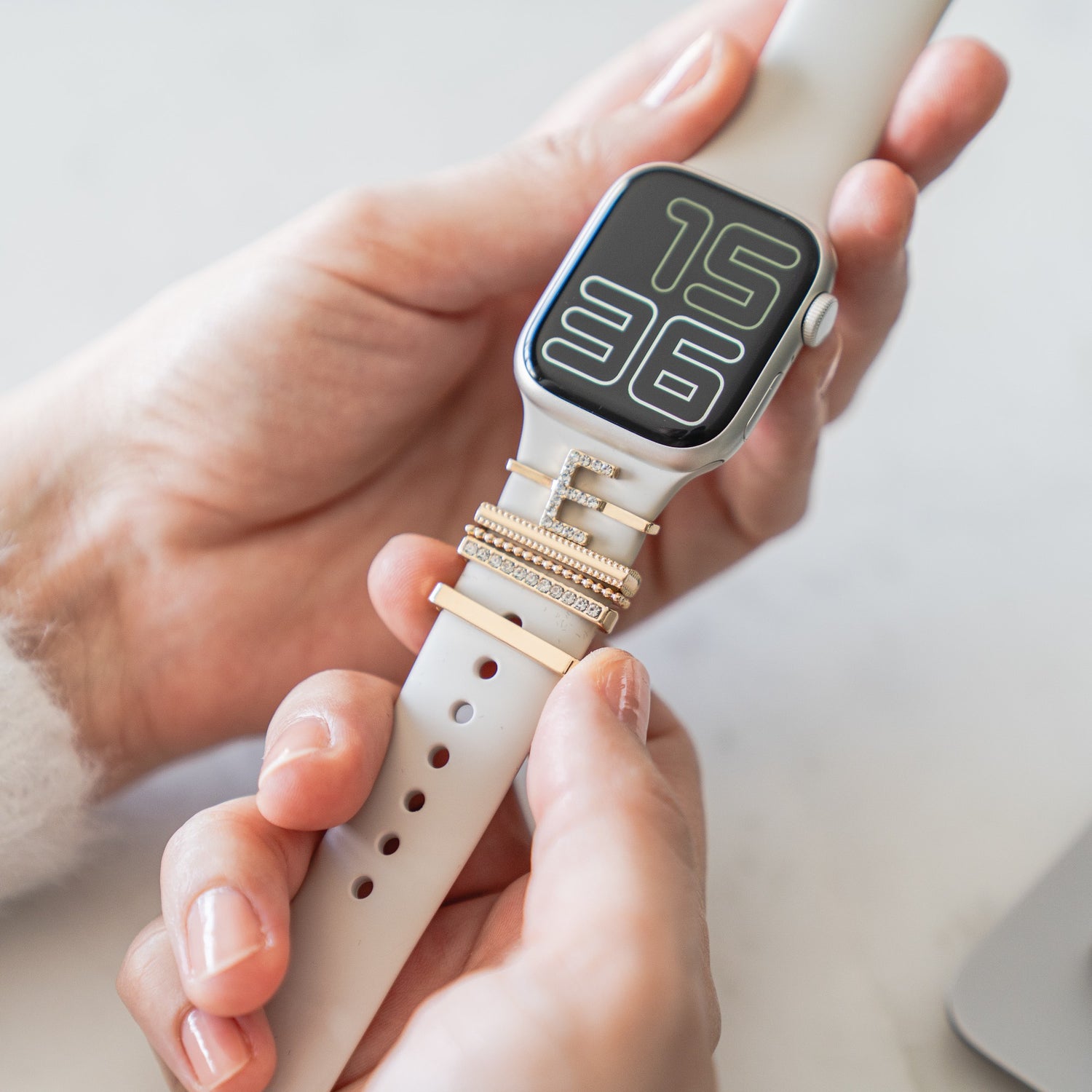 Eine Hand hält eine Apple Watch mit einem weißen Arktisband und gestapelten goldenen Ringen, die die Uhrzeit 15:36 anzeigt – gestylt mit den „E Style“-Apple-Watch-Charms von Arktisband für einen schicken Accessoire-Look.