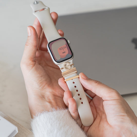 Eine Person hält eine beige Smartwatch mit goldenen Ringen am Armband neben einem geschlossenen Laptop – perfekt, um die stylischen Arktisband Apple Watch Charms „G Style“ zu präsentieren.