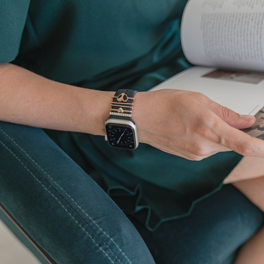 Eine Person, die eine mit goldenen „Herz“-Anhängern von Arktisband verzierte Apple Watch von Arktisband trägt, liest auf einem grünen Stuhl sitzend ein Buch.