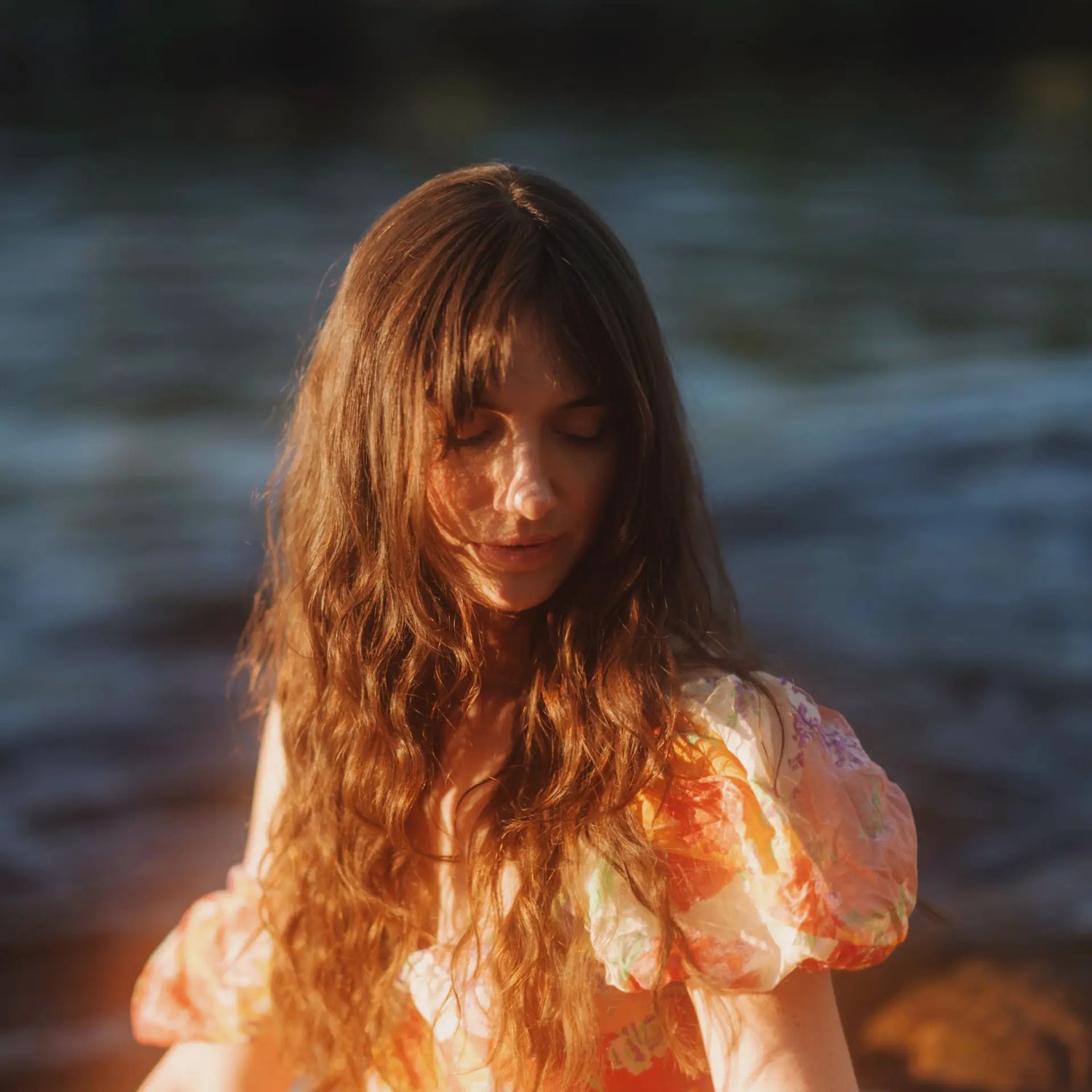 Eine Frau mit langem, gewelltem Haar in einem geblümten Kleid sitzt am Wasser und leuchtet sanft im warmen Sonnenlicht, als würde sie durch den MOMENT 67mm Variable ND (2-7 Stop) & CineBloom 10 Filter betrachtet.