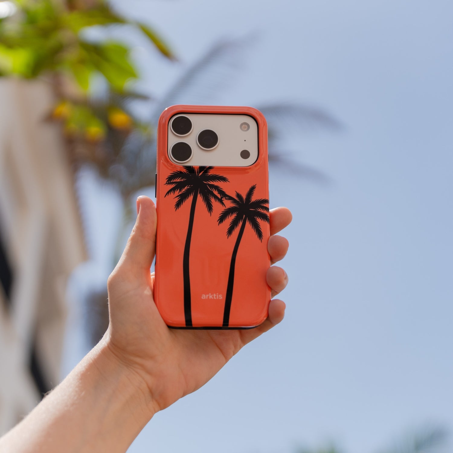 Eine Hand hält die „Arktis iPhone 18 Pro Max Hülle – Coral Coast“, eine stilvolle TPU-Silikon-Hülle von arktis mit zwei schwarzen Palmen vor einem blauen Himmel. Die Hülle ist MagSafe-kompatibel und bietet somit sowohl Schutz als auch Komfort.