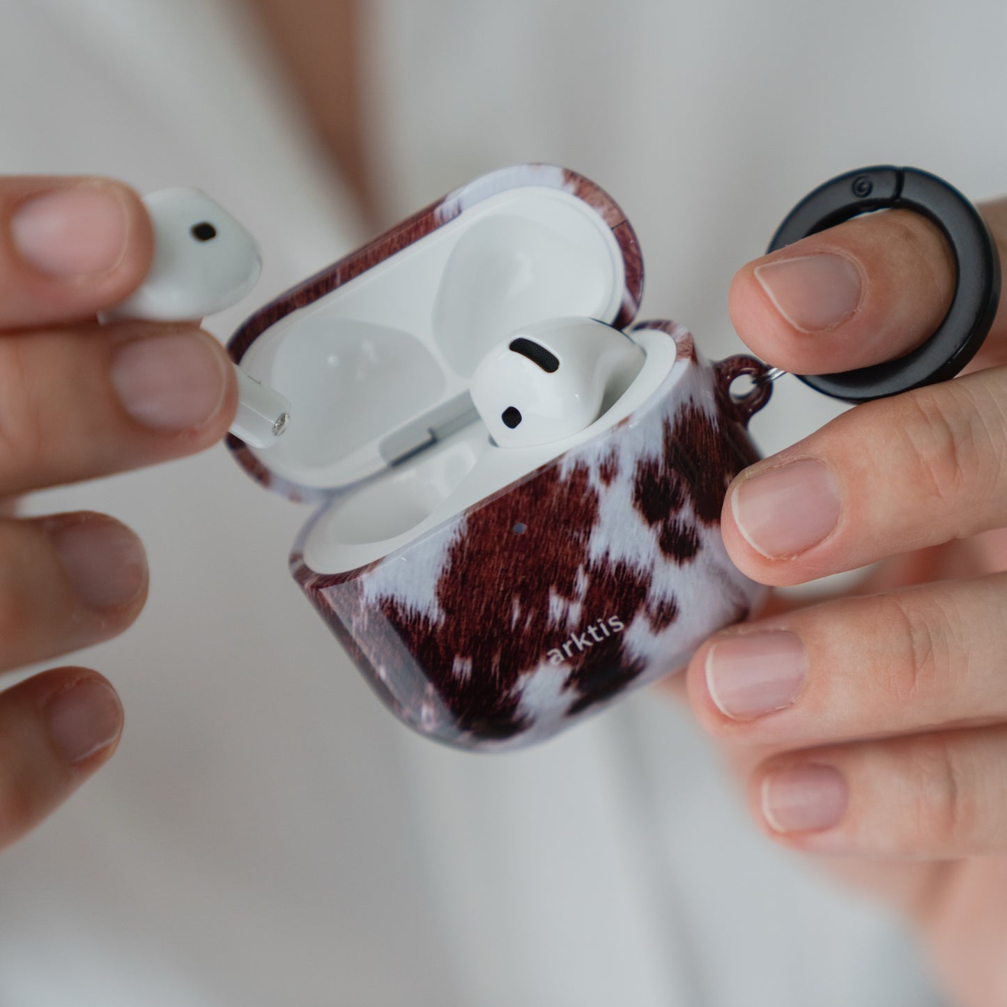 Hände, die eine geöffnete Arktis AirPods 5-Hülle – Cow Case – halten, wobei ein Ohrhörer herausgenommen wurde.