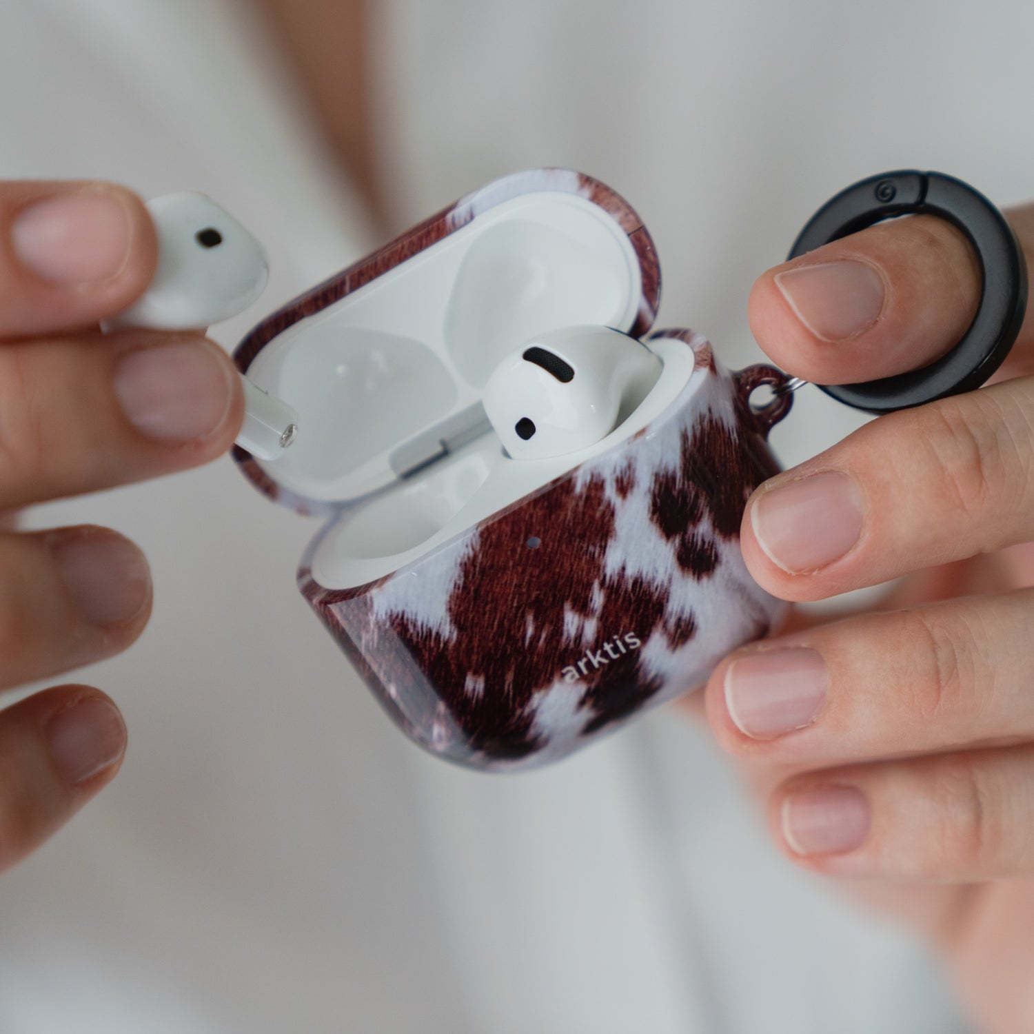 Hände, die eine geöffnete Arktis AirPods 5-Hülle – Cow Case – halten, wobei ein Ohrhörer herausgenommen wurde.