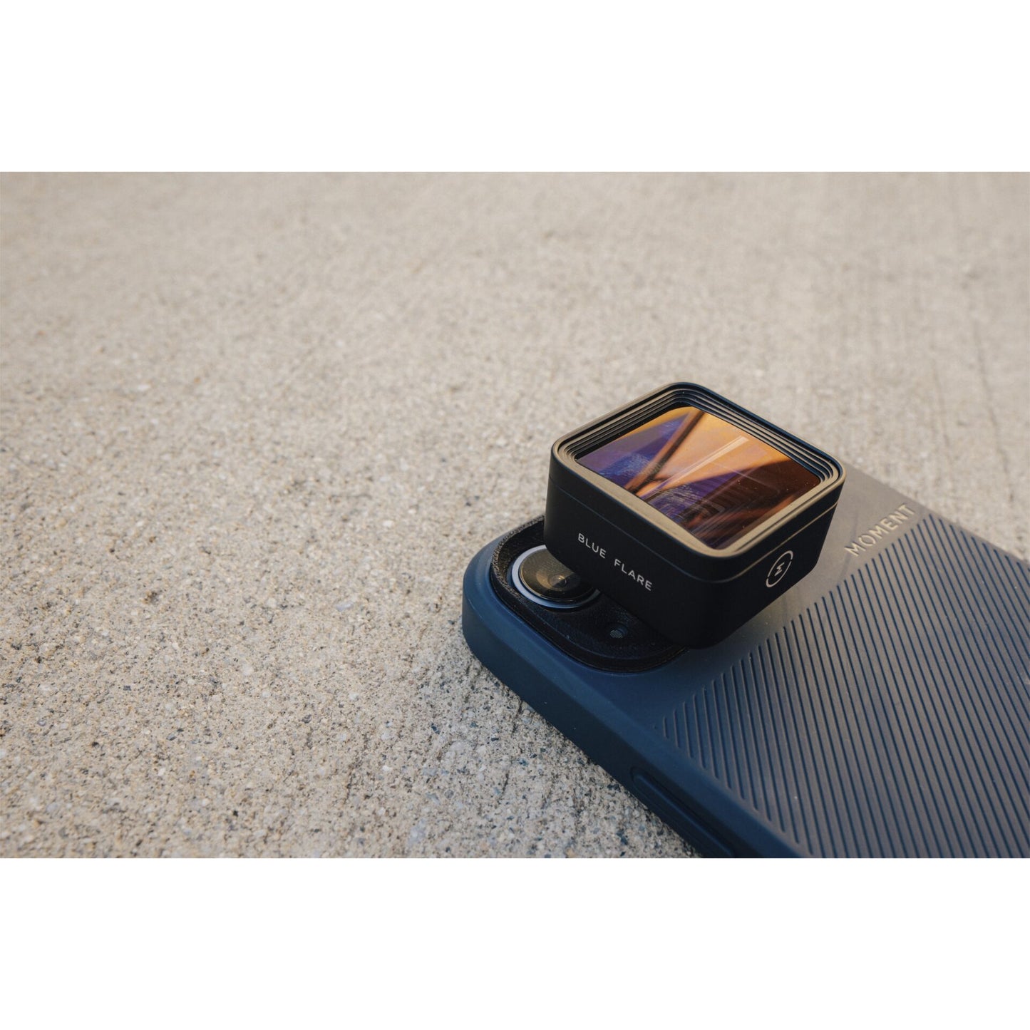 Ein Smartphone mit blauem Gehäuse und dem MOMENT 1,33x Anamorphot-Objektiv der T-Serie ruht auf einer strukturierten Betonoberfläche, bereit für kreative Smartphone-Filme.